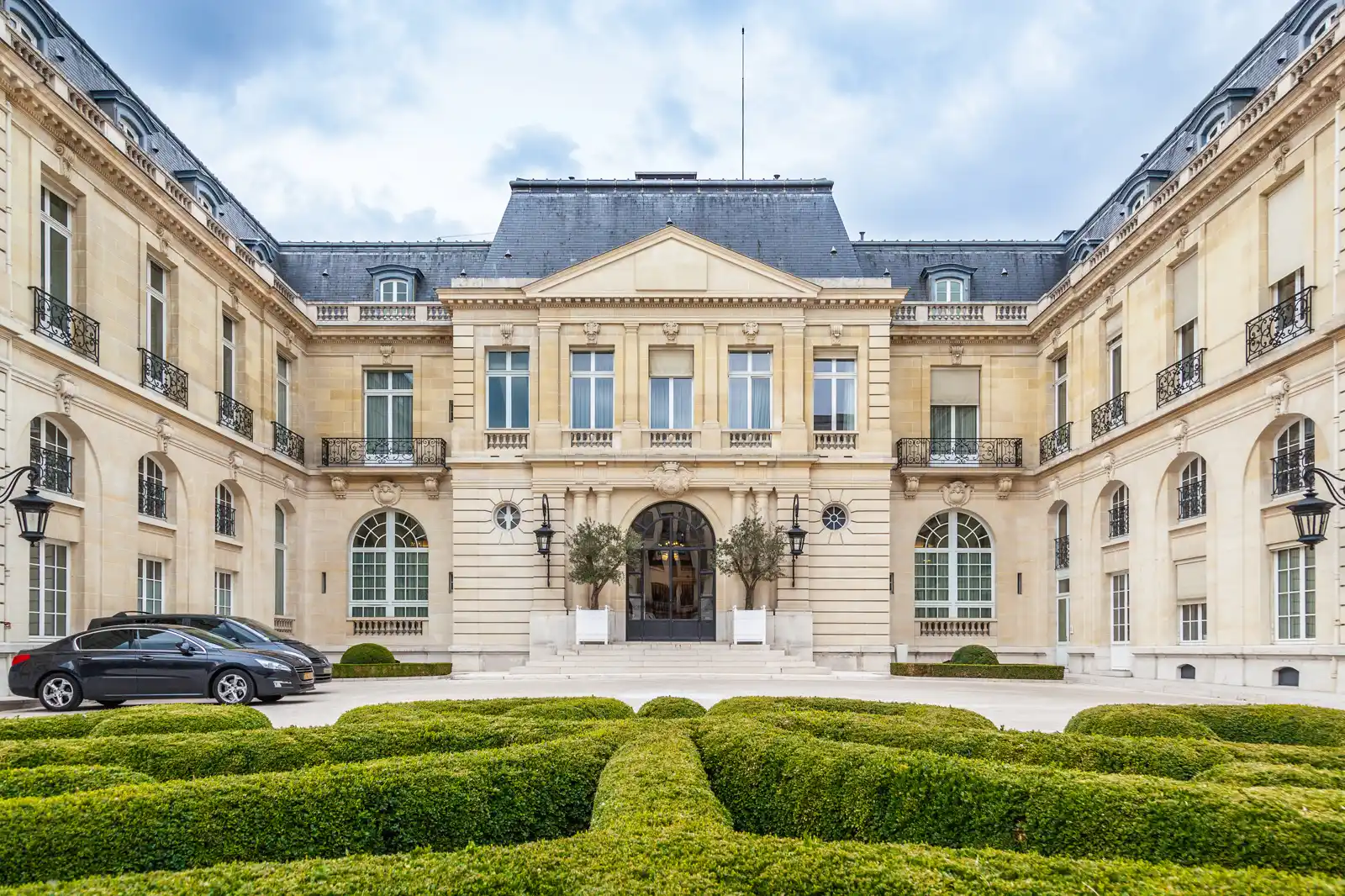 Patio de honor de un palacio de piedra clara de tres plantas con tejado de pizarra y frontón sobre la puerta principal, con las dos alas curvándose hacia el espectador; en primer plano, setos de boj recortados y un coche aparcado junto al ala izquierda.