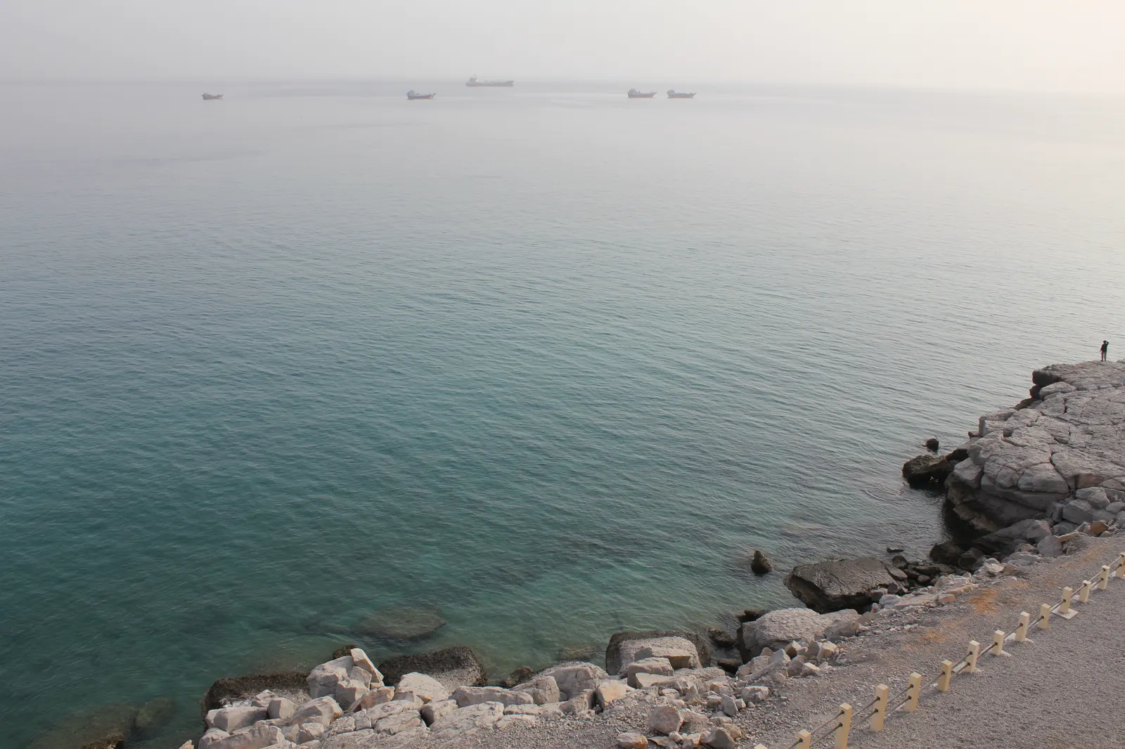 Costa rocosa y camino de grava junto a un mar liso de tonos turquesa; en el horizonte, entre la calima, se recortan las siluetas de varios cargueros fondeados.