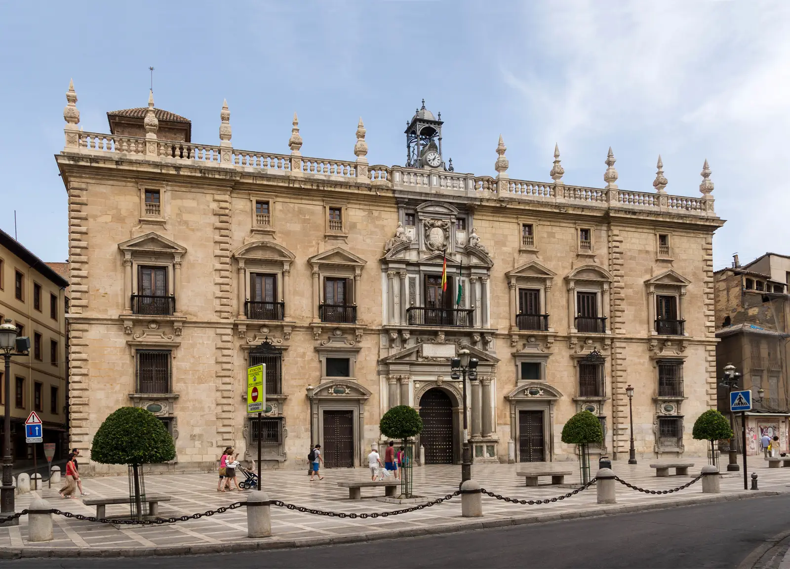 Fachada renacentista de piedra de la Real Chancillería de Granada, de dos plantas, con balcones de forja, escudo tallado sobre el balcón central, banderas de España y Andalucía, torre del reloj y viandantes cruzando la plaza empedrada delante del edificio.
