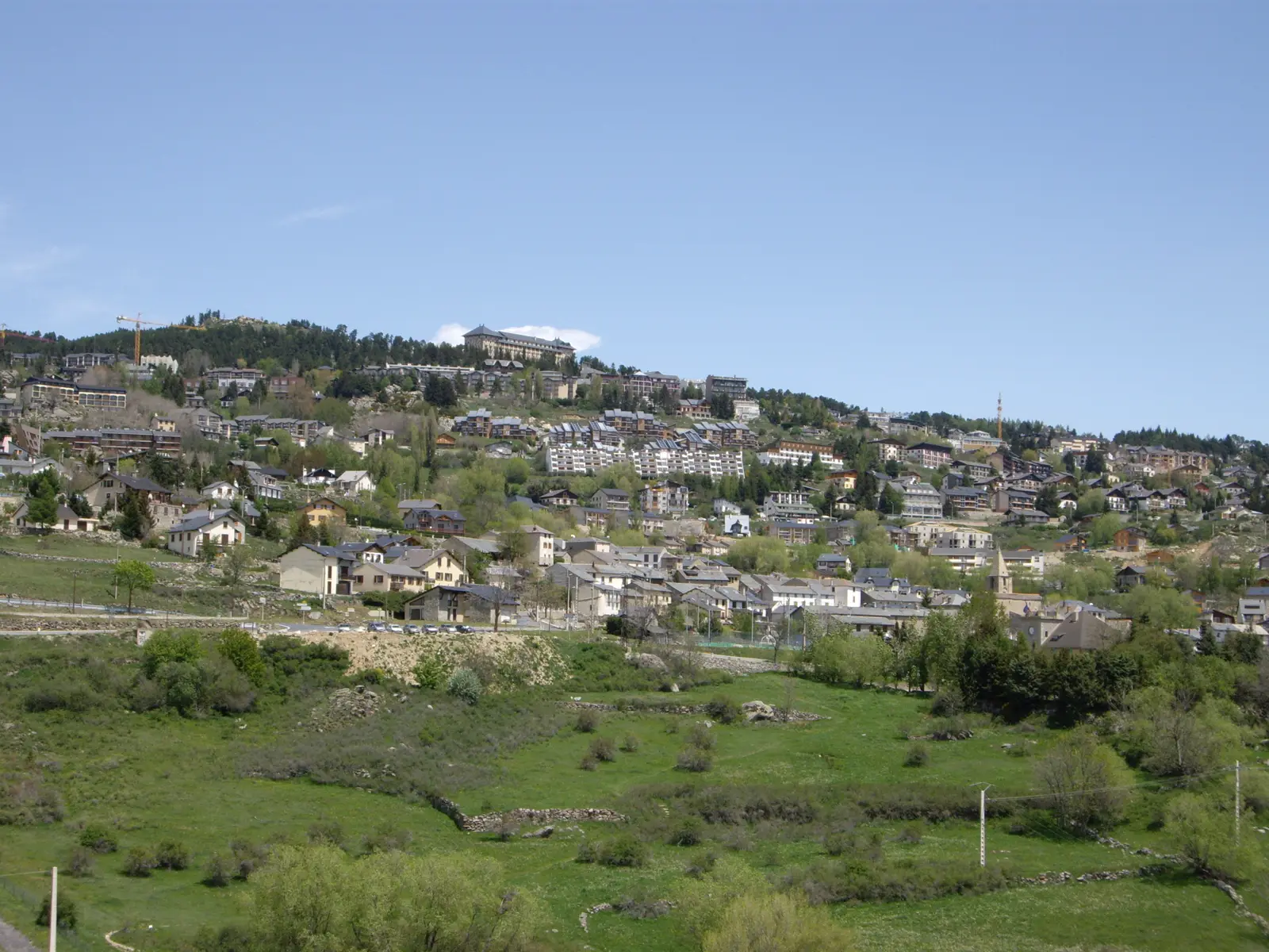 Vista general de Font-Romeu-Odeillo-Via: casas y bloques de apartamentos escalonados en una ladera boscosa de los Pirineos, con prados verdes en primer plano y cielo despejado.