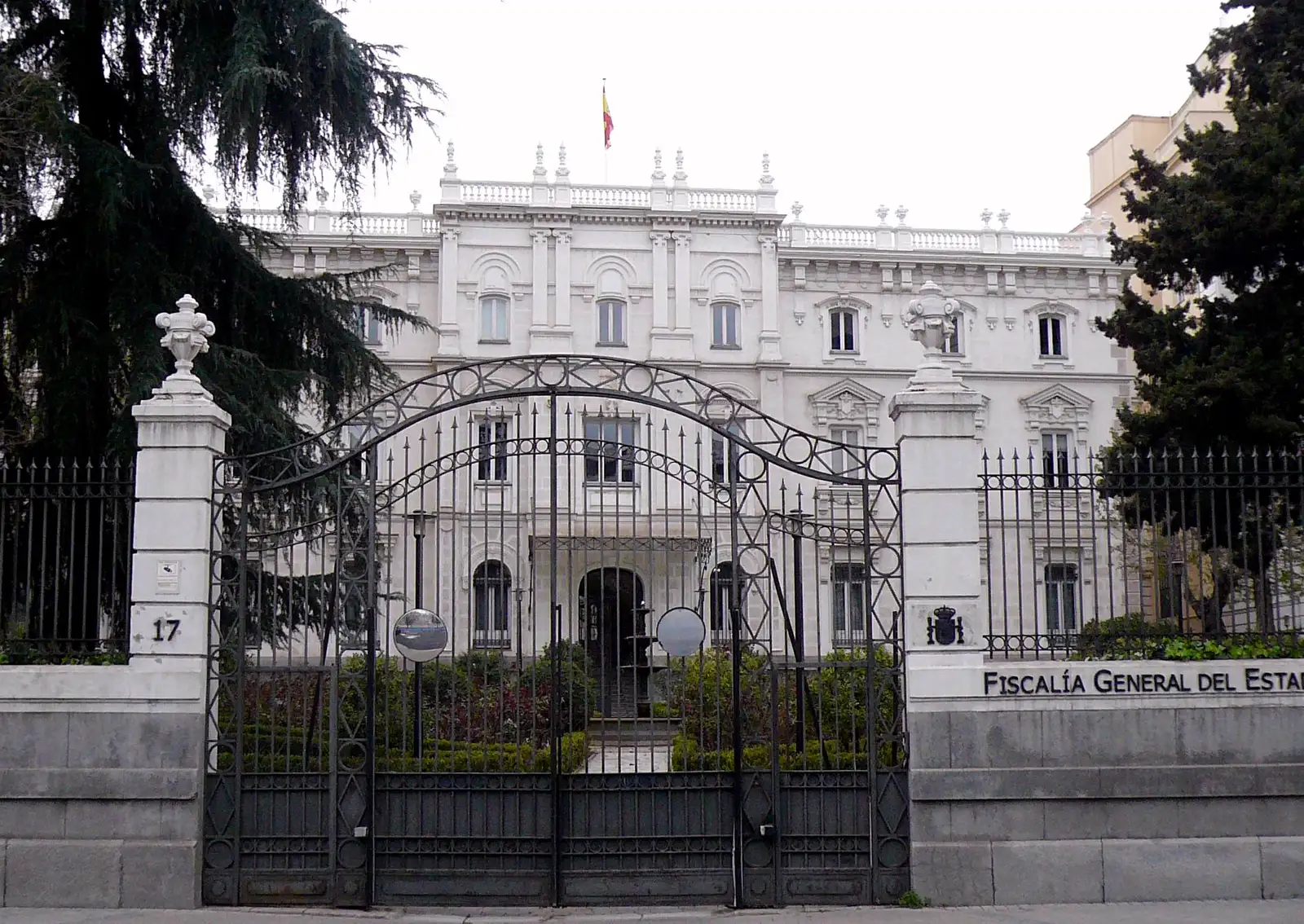 Fachada de un palacio blanco de tres plantas tras una verja de hierro cerrada, con la bandera de España en el tejado y el rótulo «Fiscalía General del Estado» sobre el muro de piedra.