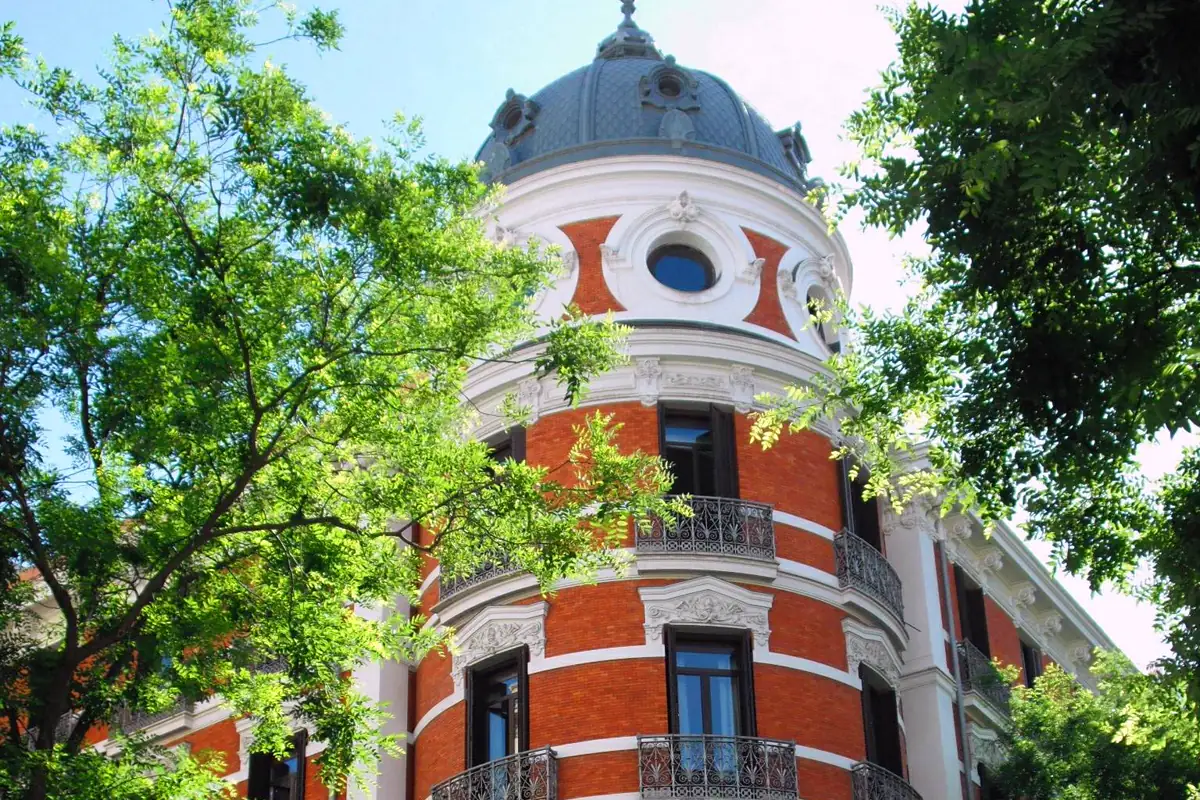 Esquina en chaflán curvo de un edificio de ladrillo rojo con molduras blancas y balcones de hierro forjado, rematada por una cúpula de pizarra con un óculo ovalado, entre las ramas de dos árboles y bajo un cielo azul.