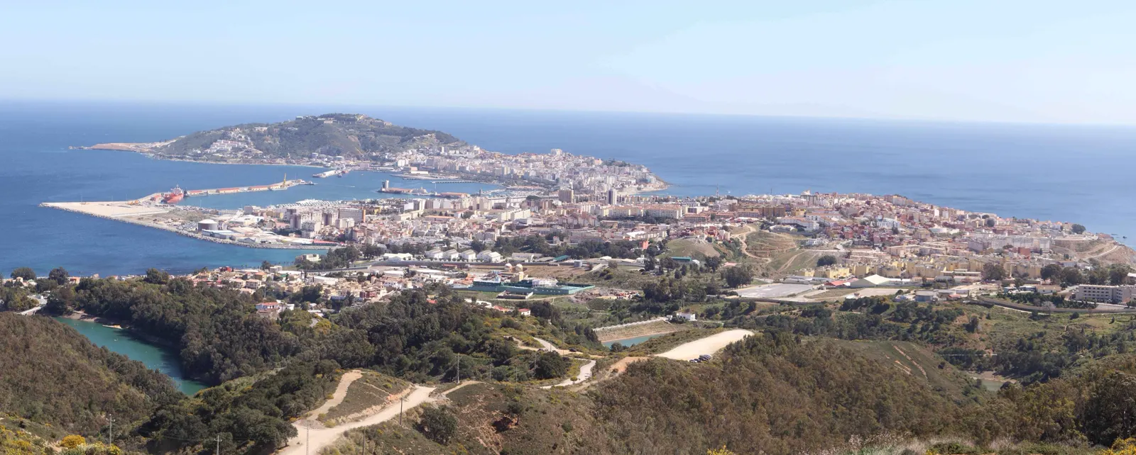 Panorámica diurna de Ceuta desde una loma: el casco urbano ocupa el istmo entre dos brazos de mar, con el puerto y sus diques a la izquierda, bloques de viviendas y naves industriales en el centro y el monte Hacho al fondo, sobre el Mediterráneo.