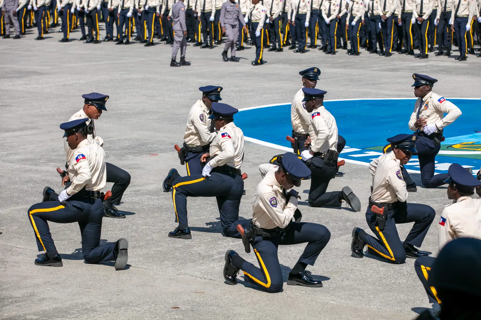 Una decena de agentes de la Policía Nacional de Haití, con camisa beis, pantalón azul de raya amarilla y gorra de plato, hincan una rodilla en el suelo de hormigón de un patio durante una ceremonia; al fondo, otras formaciones de agentes en pie.