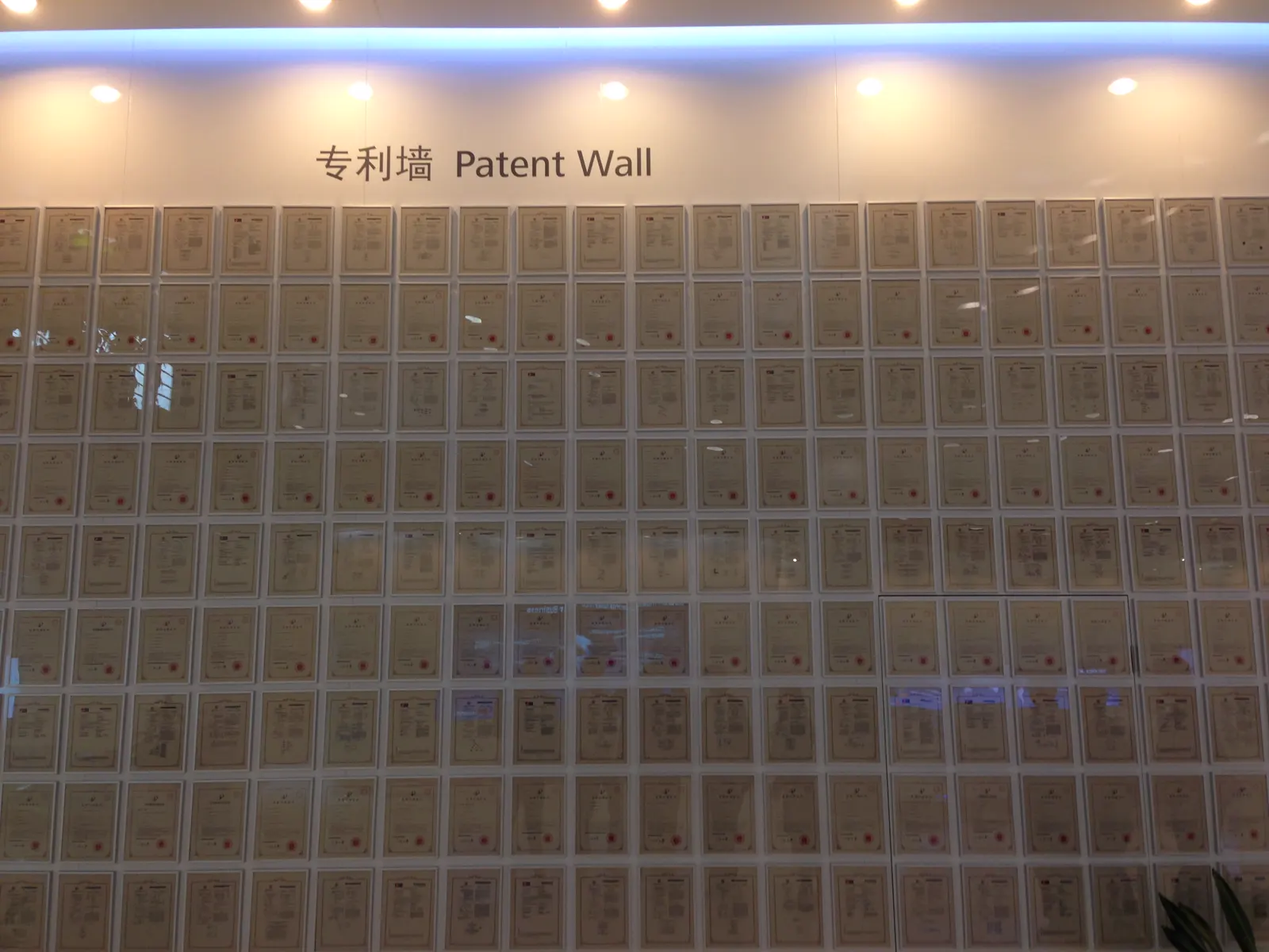 Pared cubierta de cientos de certificados de patente enmarcados, ordenados en filas bajo un rótulo en chino e inglés que dice «Patent Wall» (muro de patentes)