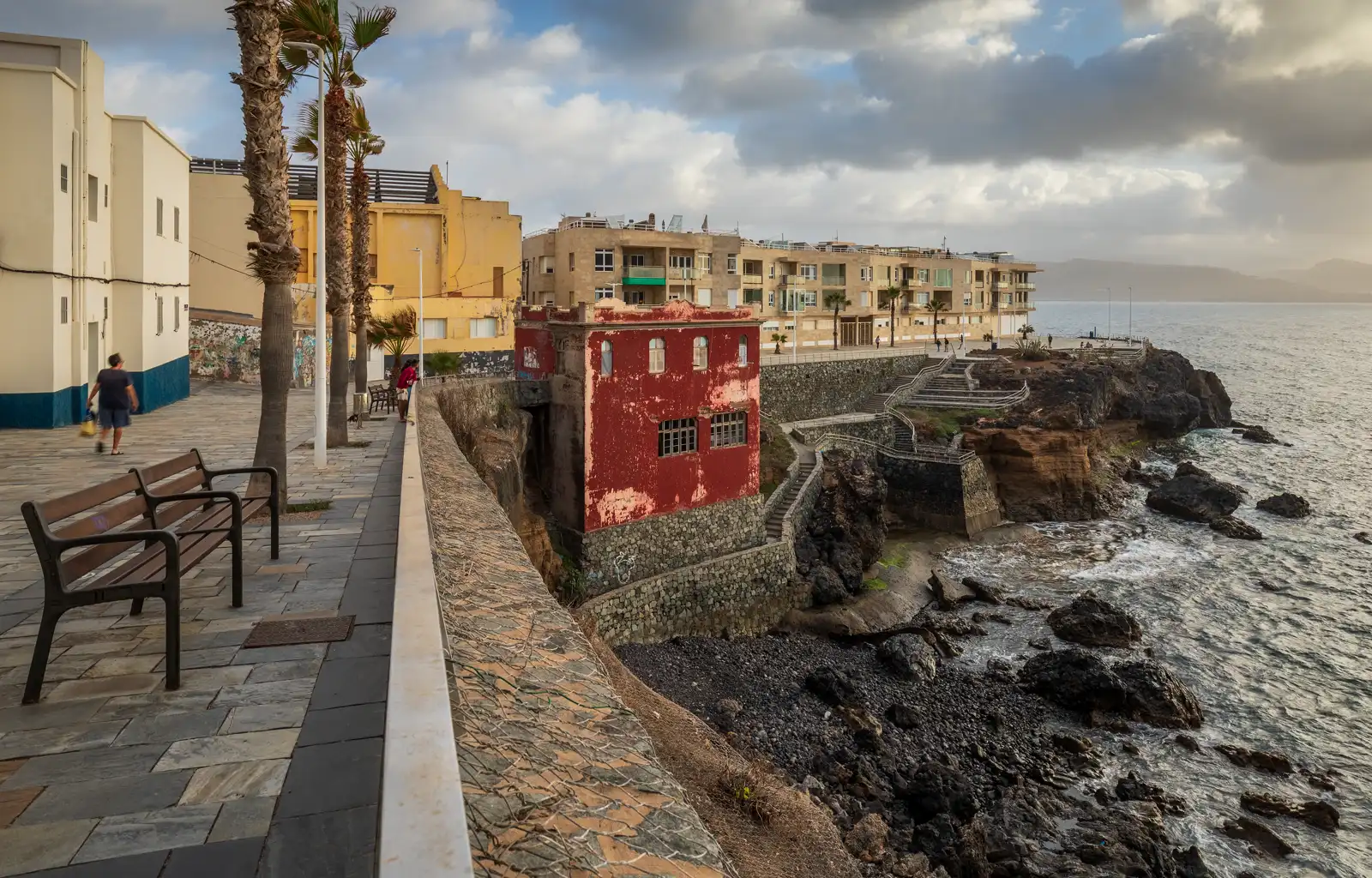 Paseo marítimo con bancos y palmeras junto a bloques de viviendas de varias alturas asomados a una costa rocosa, con el mar y un cielo nublado al fondo.