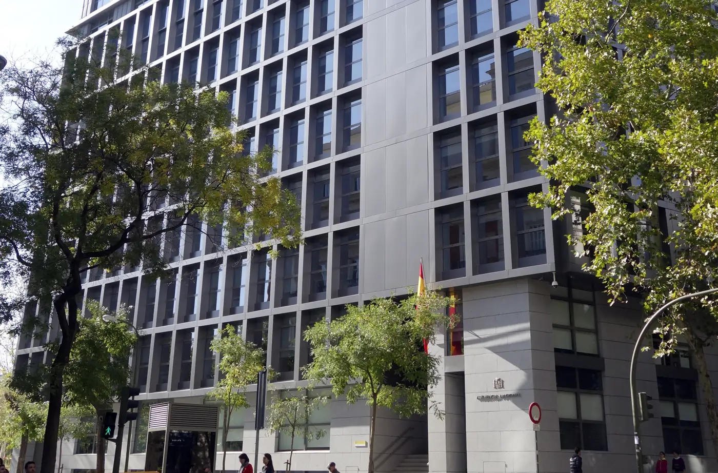 Fachada del edificio de la Audiencia Nacional en Madrid, un bloque moderno de varias plantas con ventanas rectangulares enmarcadas en gris; junto a la puerta principal, el escudo de España y el rótulo «Audiencia Nacional», con árboles y viandantes en la acera.