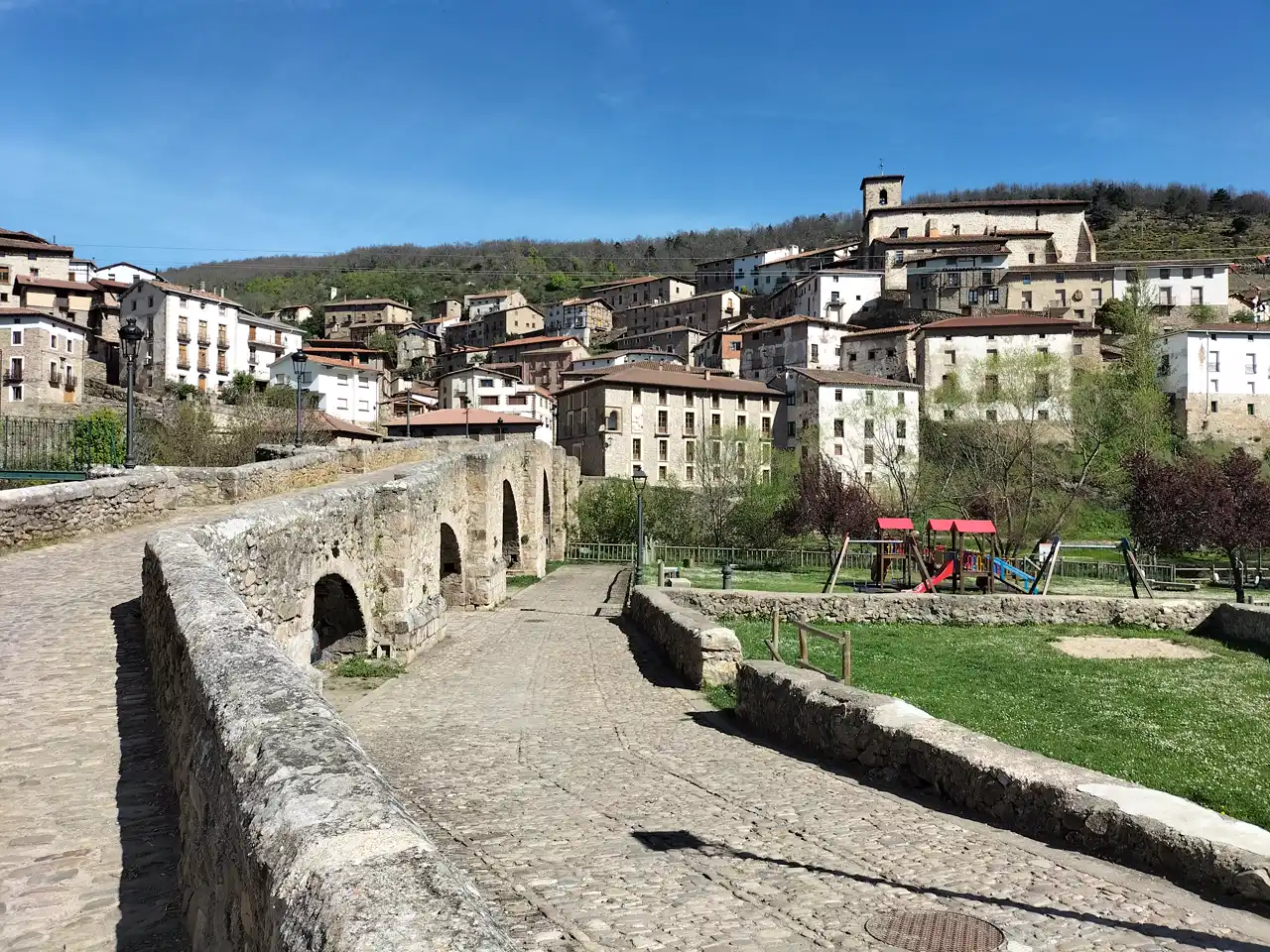 Villoslada de Cameros visto desde su puente medieval de piedra: casas de sillar y tejado rojo trepando por la ladera, la torre de la iglesia recortada arriba a la derecha y, detrás del caserío, una loma cubierta de arbolado. En primer plano, el pretil del puente y una calzada empedrada; a la derecha, un parque infantil sobre césped.