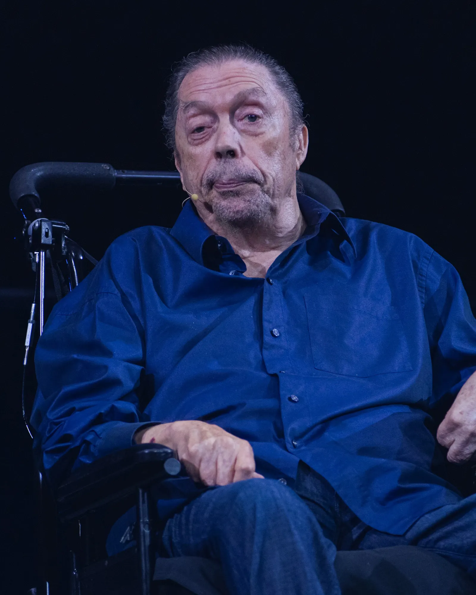 Tim Curry, hombre mayor de pelo cano recogido y barba corta, sentado en una silla de ruedas sobre un escenario a oscuras. Viste camisa azul de botones y lleva un micrófono de diadema junto a la comisura de la boca; apoya la mano derecha en el reposabrazos y mira hacia el público.