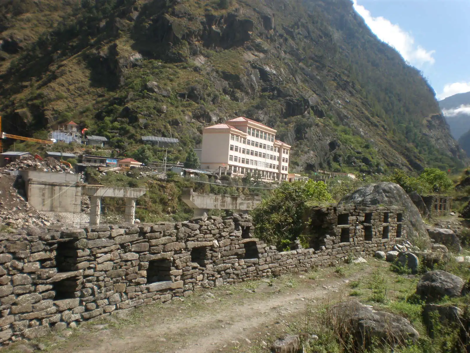 Fortaleza de piedra en ruinas sobre una ladera boscosa junto a un río encajonado entre montañas; al otro lado del cauce se ven los edificios blancos del paso fronterizo chino de Gyirong