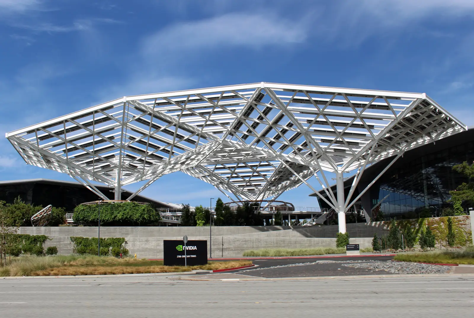 Edificio acristalado de la sede de Nvidia bajo una gran marquesina blanca de celosía triangular cubierta de placas solares, con un cartel negro con el logotipo de la compañía junto a la carretera
