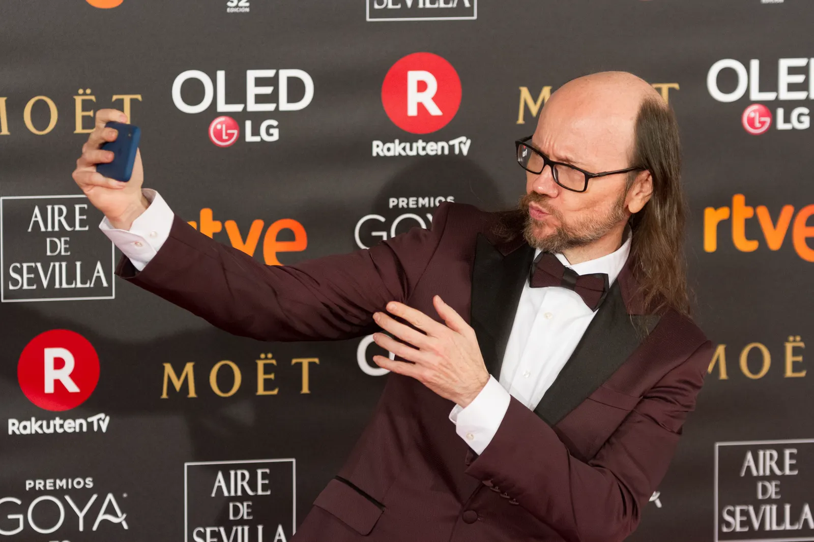 Santiago Segura, con esmoquin granate y pajarita, se hace un selfi con el móvil ante el panel de patrocinadores de los Premios Goya.