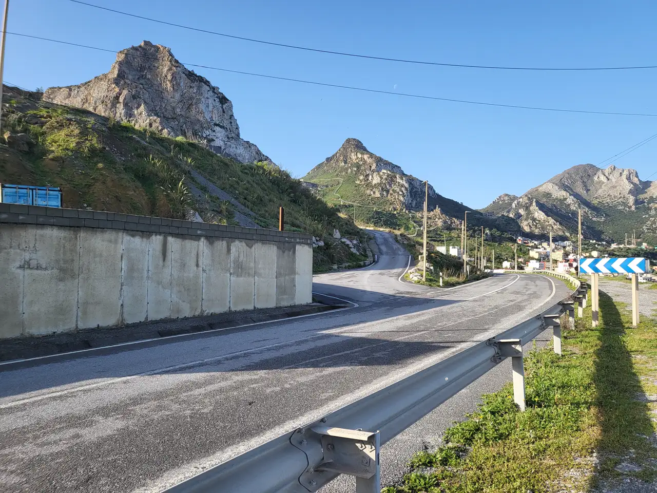 Carretera de dos carriles con quitamiedos y muro de contención que discurre entre montañas rocosas hacia el caserío de Benzú, en Ceuta, bajo un cielo despejado.