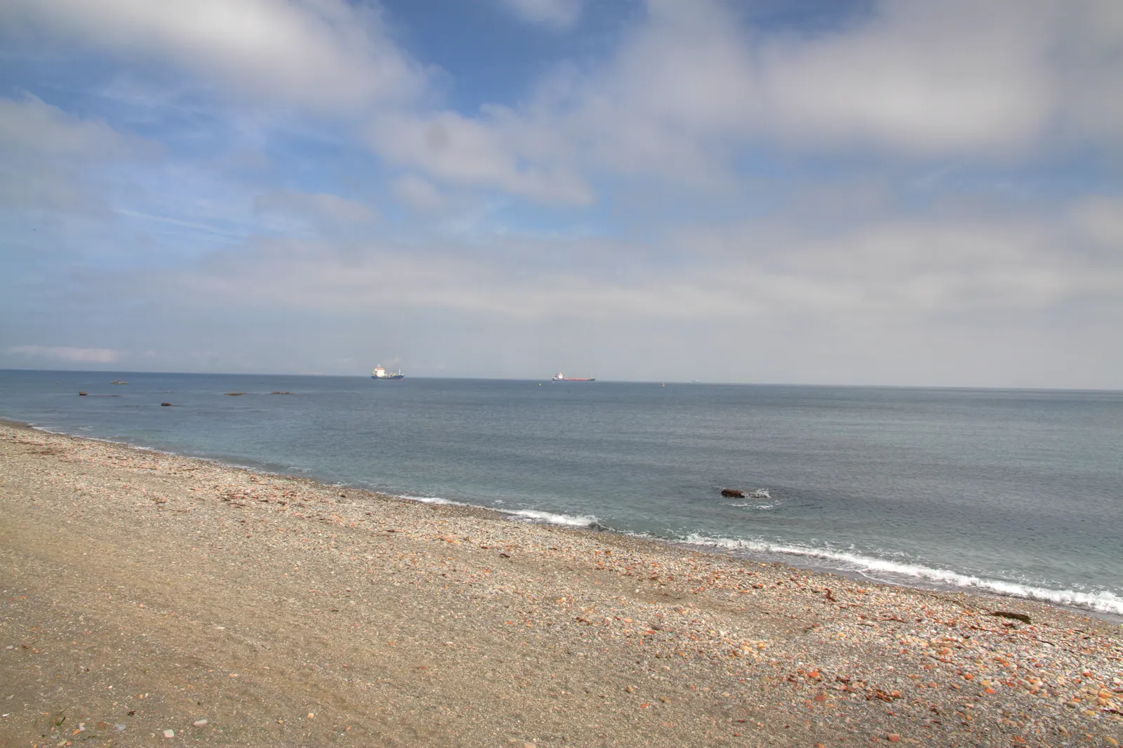 Playa de arena y grava vacía junto a un mar en calma, con dos buques fondeados en el horizonte bajo un cielo con nubes altas.