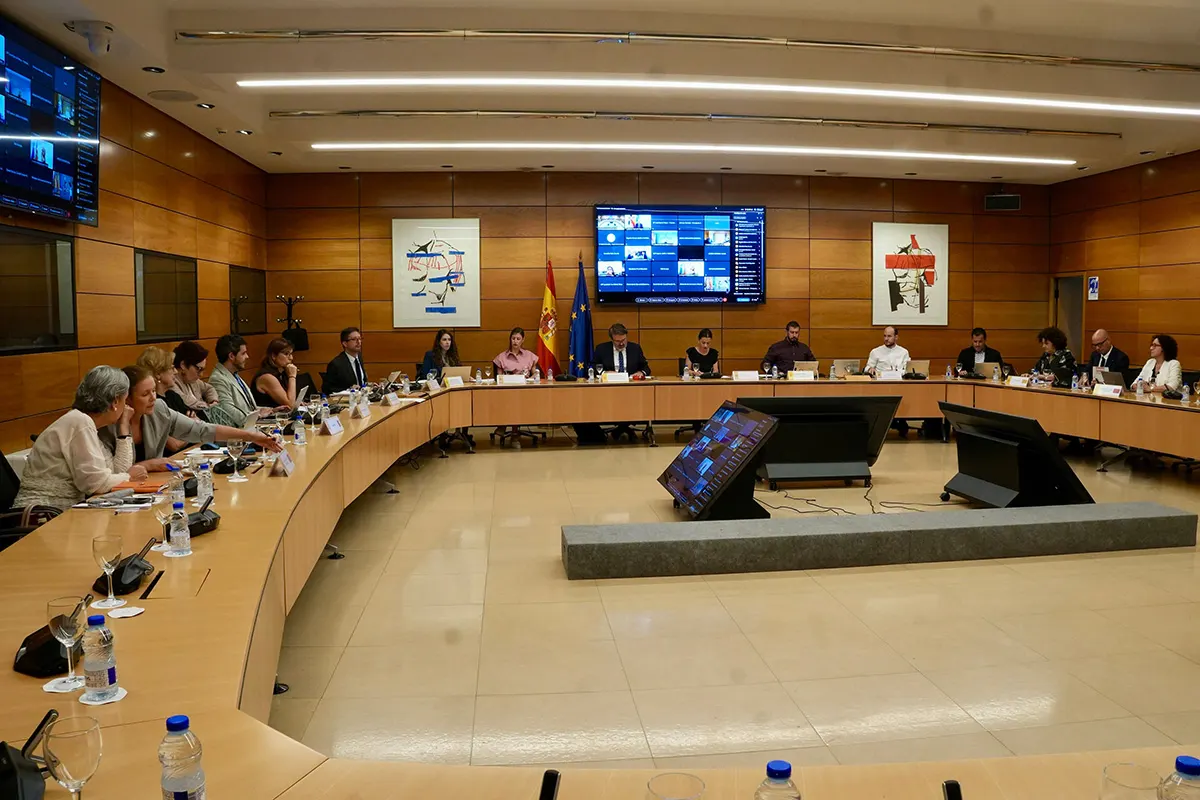 Sala de reuniones con paredes de madera y una gran mesa en herradura ocupada por una quincena de personas con carpetas y micrófonos; al fondo, las banderas de España y de la Unión Europea junto a una pantalla que muestra en mosaico a los participantes conectados por videoconferencia.