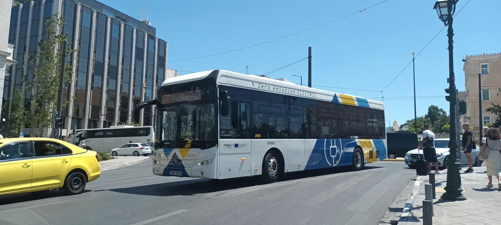 Autobús urbano eléctrico blanco con franjas azules y amarillas y el rótulo «Zero emission ELECTRIC» en el costado, circulando por una avenida del centro de Atenas junto a un taxi amarillo, con edificios de oficinas y peatones en la acera.