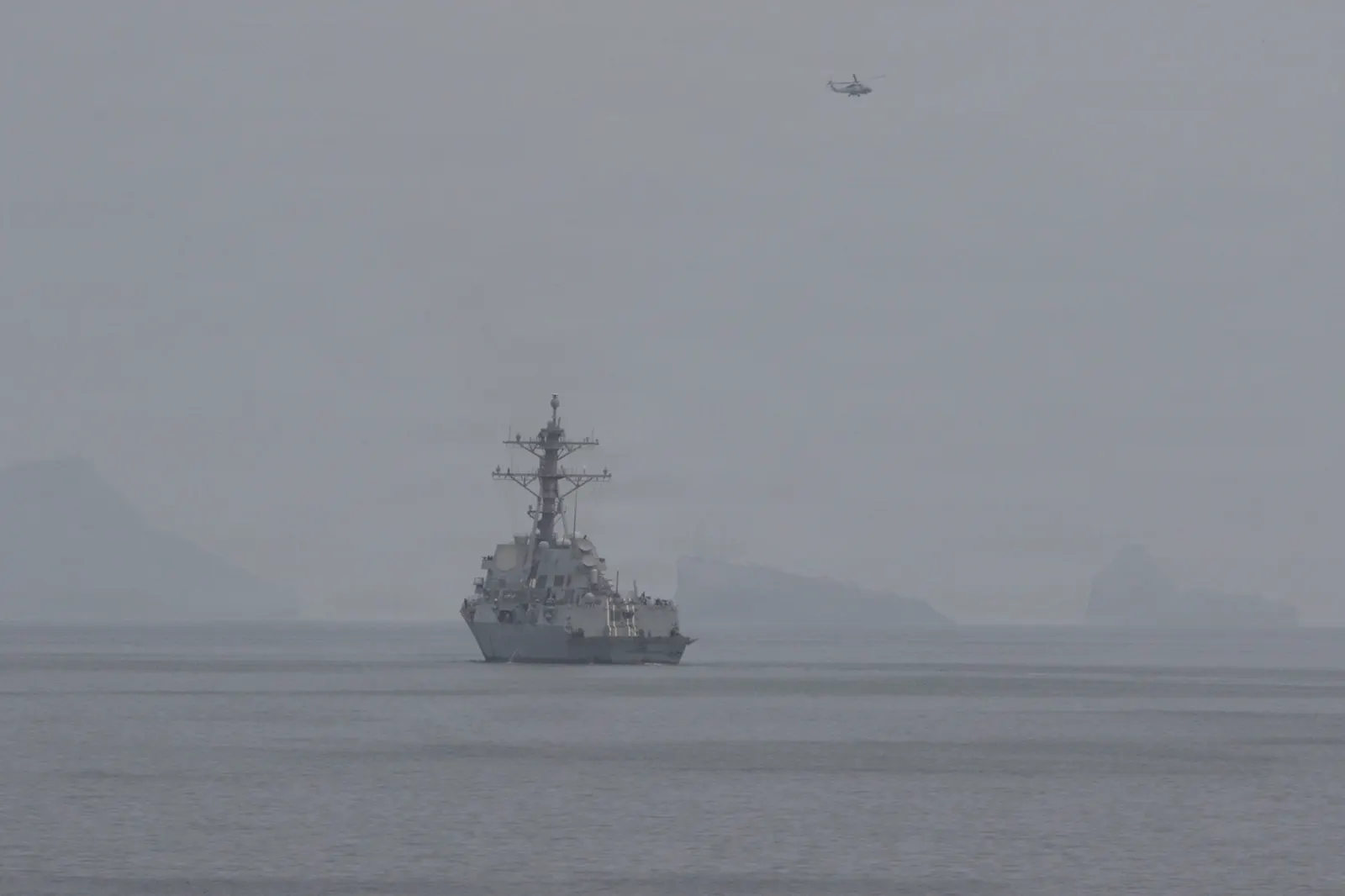 Un destructor gris de la Armada estadounidense navega solo por un mar en calma bajo una calima densa que borra el horizonte; sobre él vuela un helicóptero y, al fondo, se adivinan las siluetas de montañas costeras.