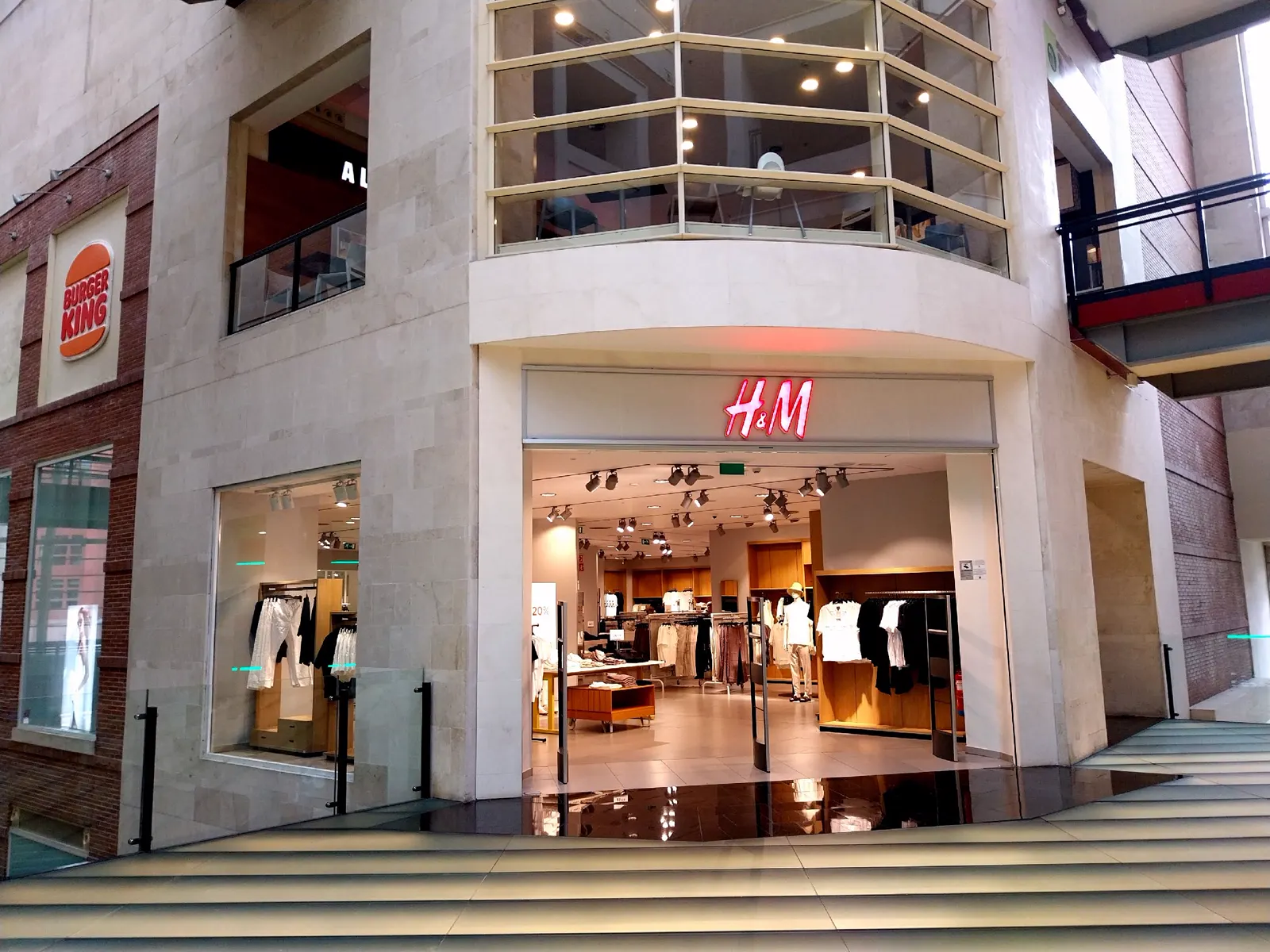Fachada de una tienda de H&M en el interior de un centro comercial, con el rótulo rojo iluminado sobre la entrada abierta y ropa colgada en percheros al fondo; a la izquierda, el logotipo de un Burger King.