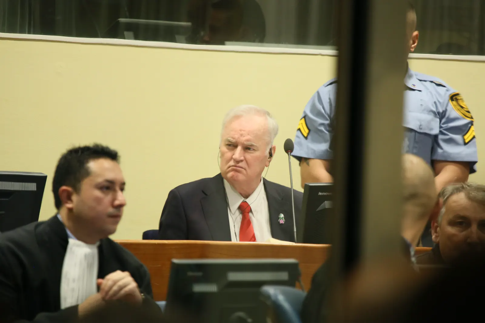 Ratko Mladic, anciano de pelo blanco y traje oscuro con corbata roja, sentado en el banquillo de una sala del tribunal de La Haya entre un abogado con toga y un agente uniformado