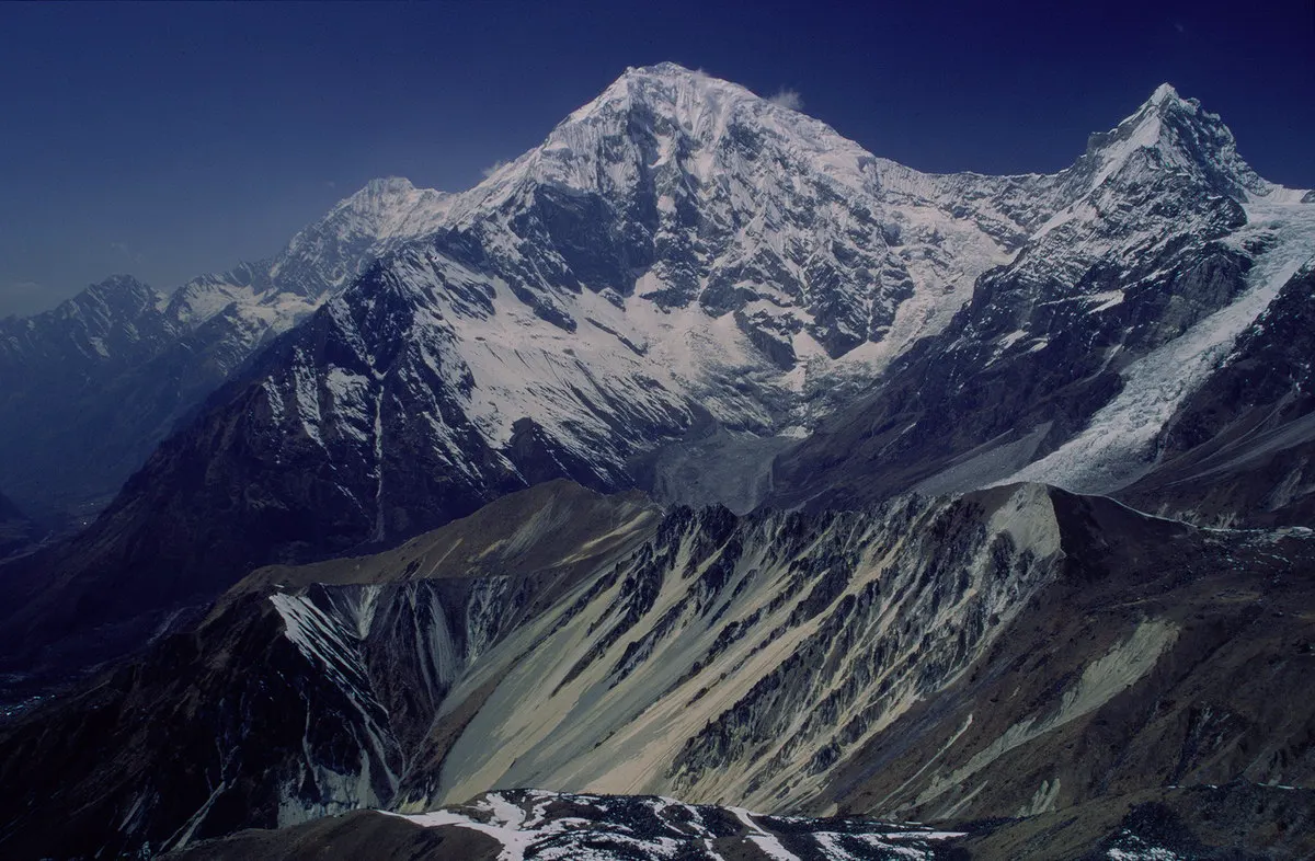 Cumbre nevada del Langtang Lirung bajo un cielo azul intenso, con una lengua de glaciar descendiendo hacia un valle de morrenas grises y laderas pardas en primer plano