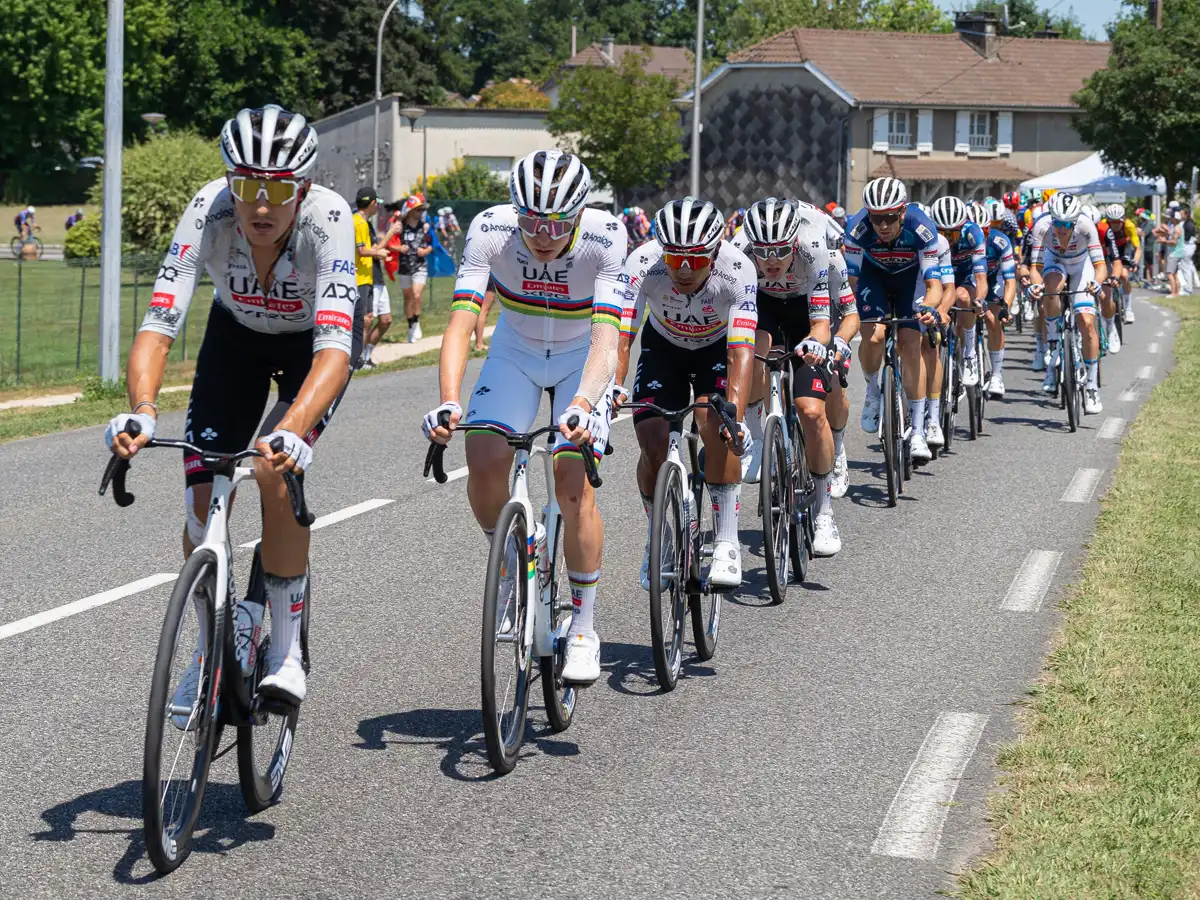 Fila de ciclistas del UAE Team Emirates encabezando el pelotón por una carretera con casas al fondo; el segundo de la fila viste el maillot blanco con franjas de arco iris de campeón del mundo.