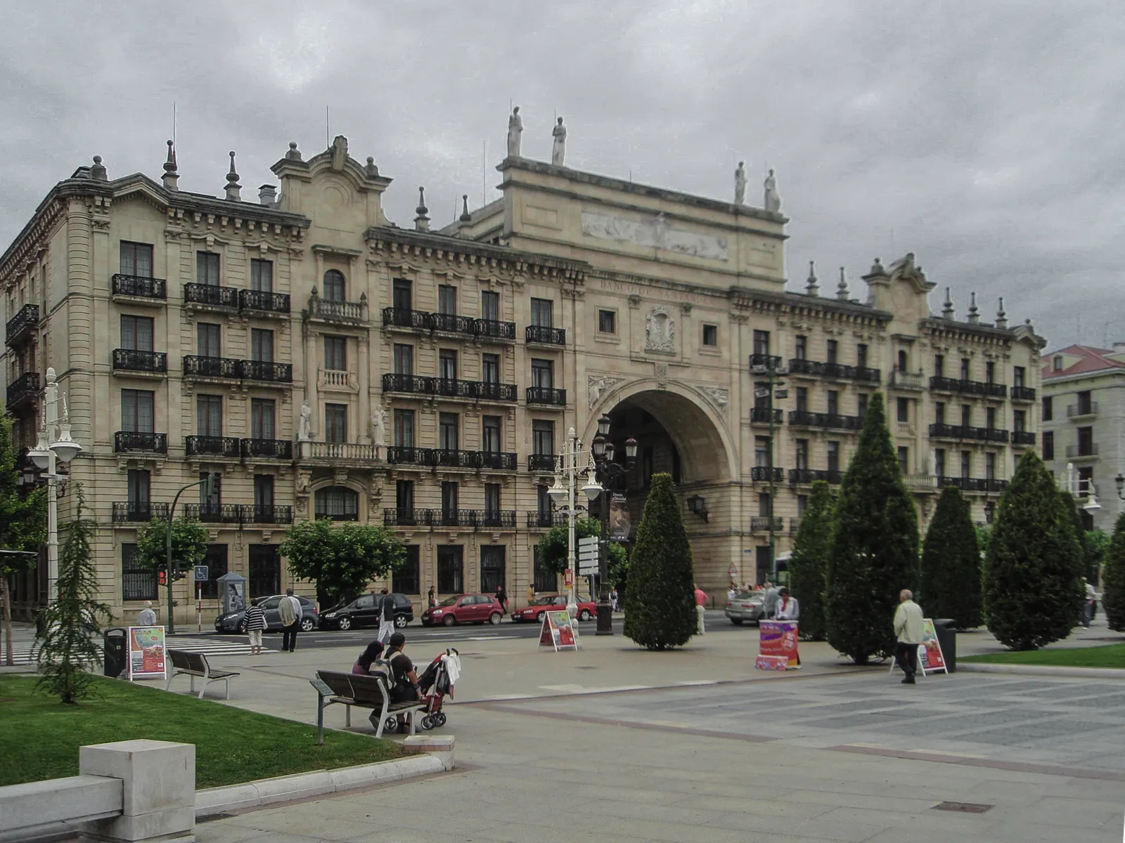 Edificio historicista de piedra clara del Banco de Santander en el Paseo de Pereda, con un gran arco central, balcones de hierro y estatuas sobre la cornisa, ante una plaza con setos recortados y transeúntes.