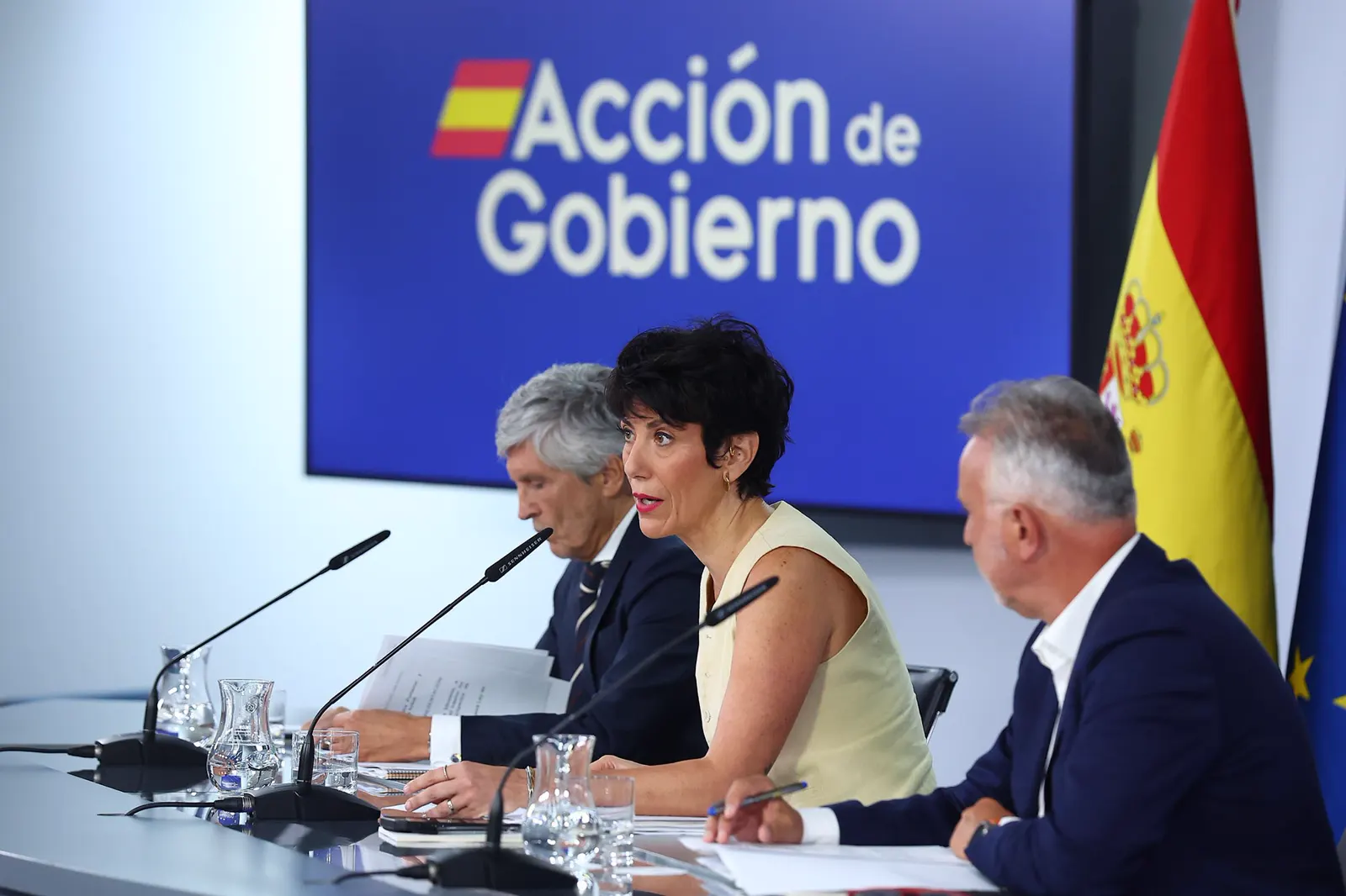 Tres ministros sentados tras una mesa con micrófonos en la sala de prensa de La Moncloa: a la izquierda, Fernando Grande-Marlaska, de pelo cano y traje azul, con la mirada baja sobre unos papeles; en el centro, Elma Saiz habla ante el micrófono; a la derecha, Ángel Víctor Torres toma notas. Al fondo, un panel azul con el rótulo «Acción de Gobierno» y las banderas de España y de la Unión Europea.