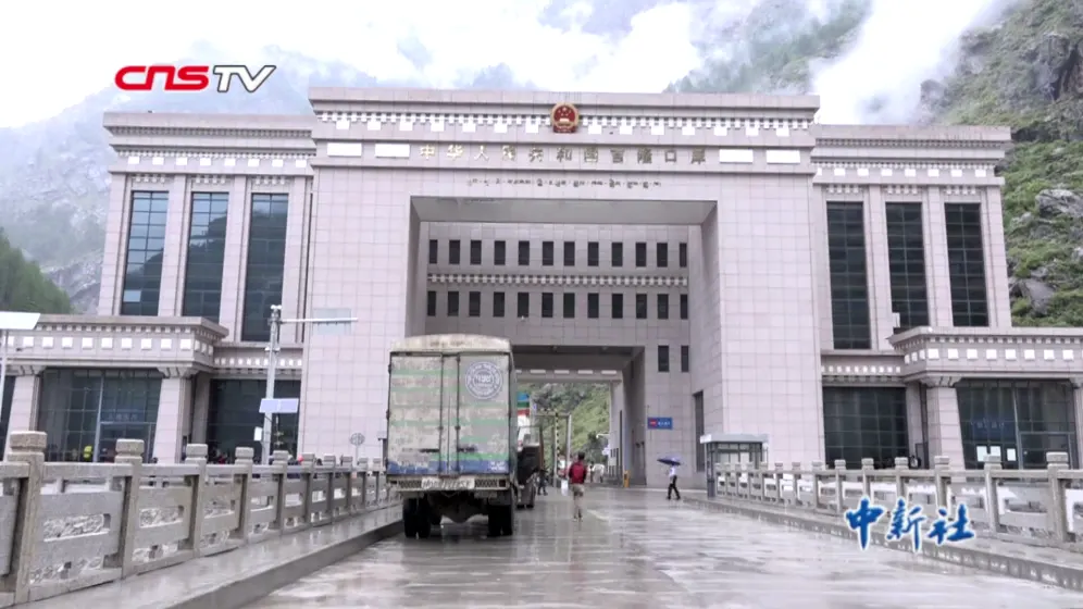 Edificio monumental del puesto fronterizo chino de Gyirong, de piedra clara y con el escudo de la República Popular sobre el arco central, con un camión y varios peatones cruzando bajo la lluvia y laderas de montaña al fondo