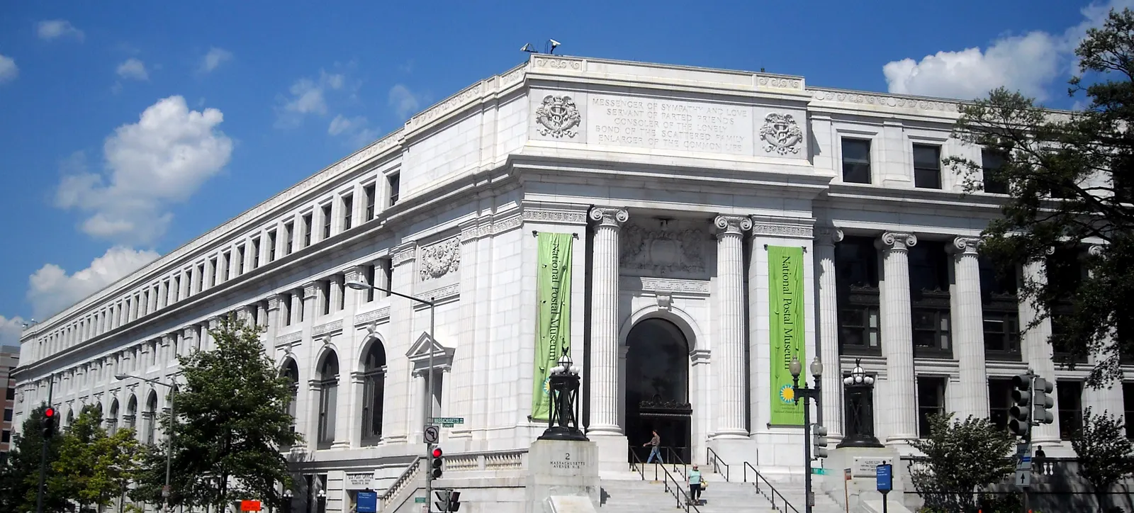Edificio neoclásico de mármol blanco con columnas y dos pancartas verdes del Museo Postal Nacional junto a la entrada, bajo cielo azul