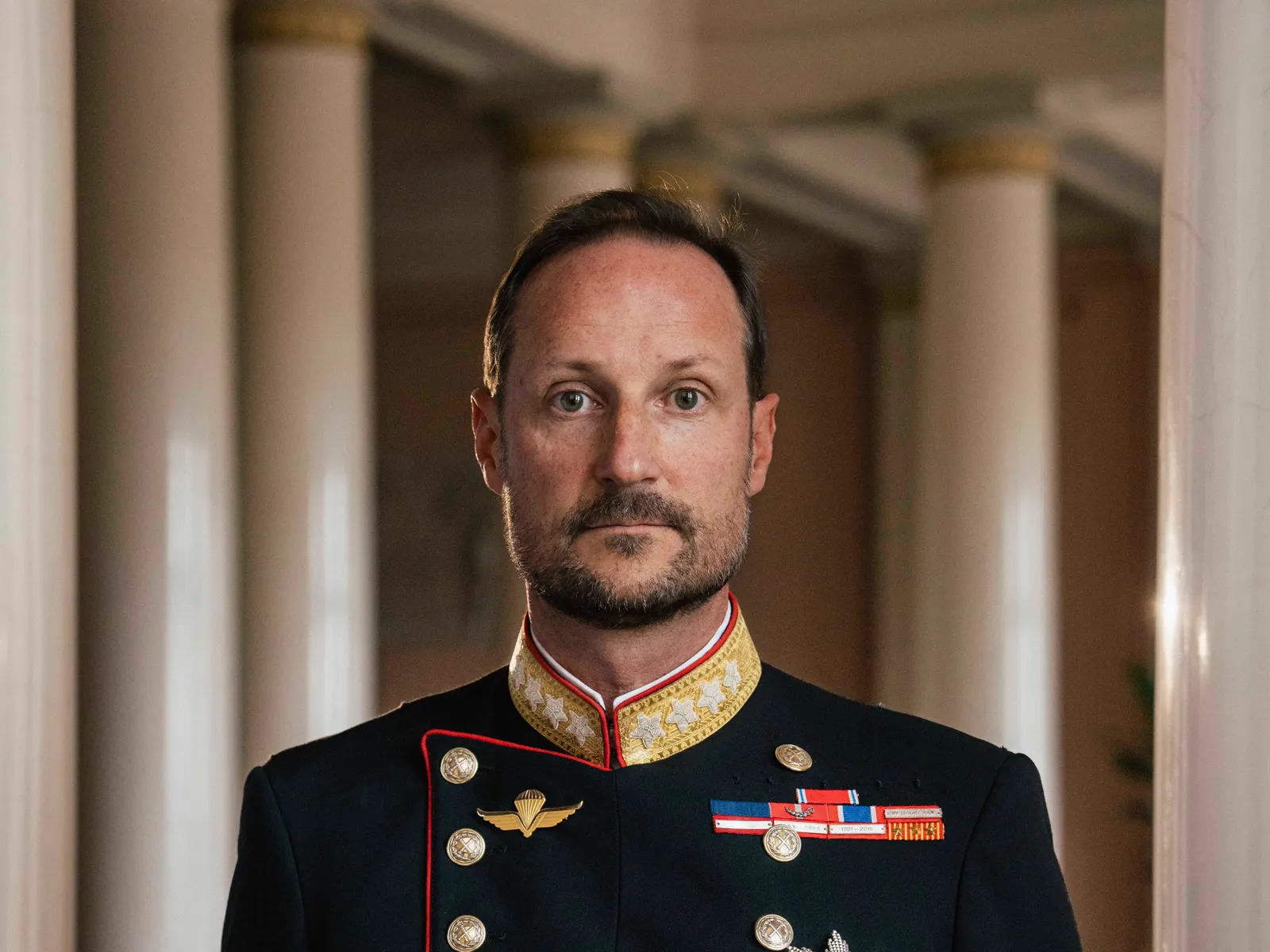 Retrato de Haakon VIII de Noruega, de pie y serio, con uniforme militar de gala verde oscuro de cuello alto bordado en oro, condecoraciones en el pecho y una gran estrella de orden, en una galería de columnas del Palacio Real de Oslo.