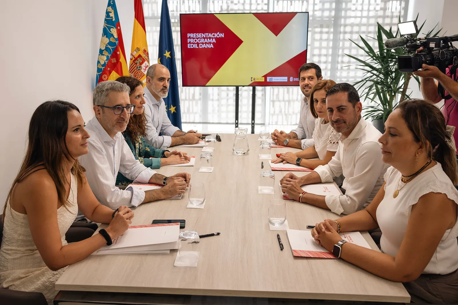 Nueve personas sentadas a ambos lados de una mesa de reuniones alargada; al fondo, las banderas de la Comunitat Valenciana, España y la Unión Europea y una pantalla con el rótulo «Presentación programa EDIL DANA». A la derecha, un cámara de televisión graba la escena.