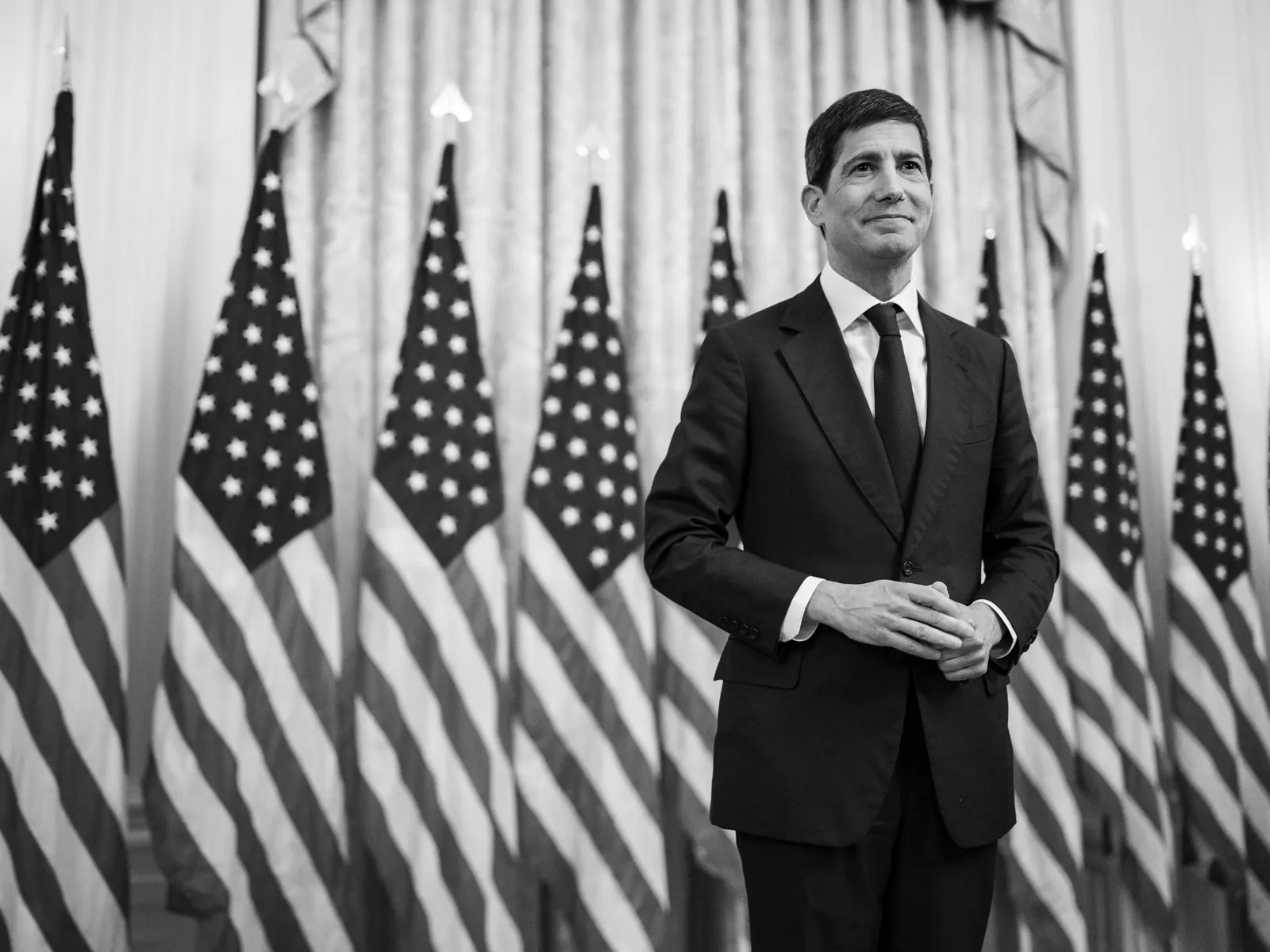 Fotografía en blanco y negro de Kevin Warsh, con traje oscuro y corbata, de pie y con las manos entrelazadas ante una hilera de banderas de Estados Unidos y cortinas de una sala oficial.