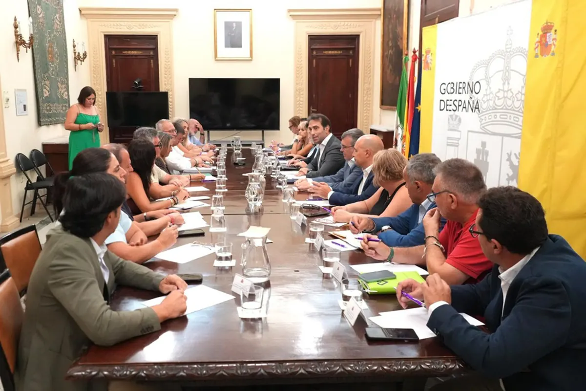 Carlos Cuerpo, sentado en el centro de una larga mesa de reuniones, escucha a los alcaldes granadinos sentados a ambos lados; al fondo, las banderas de España, Andalucía y la Unión Europea y un cartel del Gobierno de España.