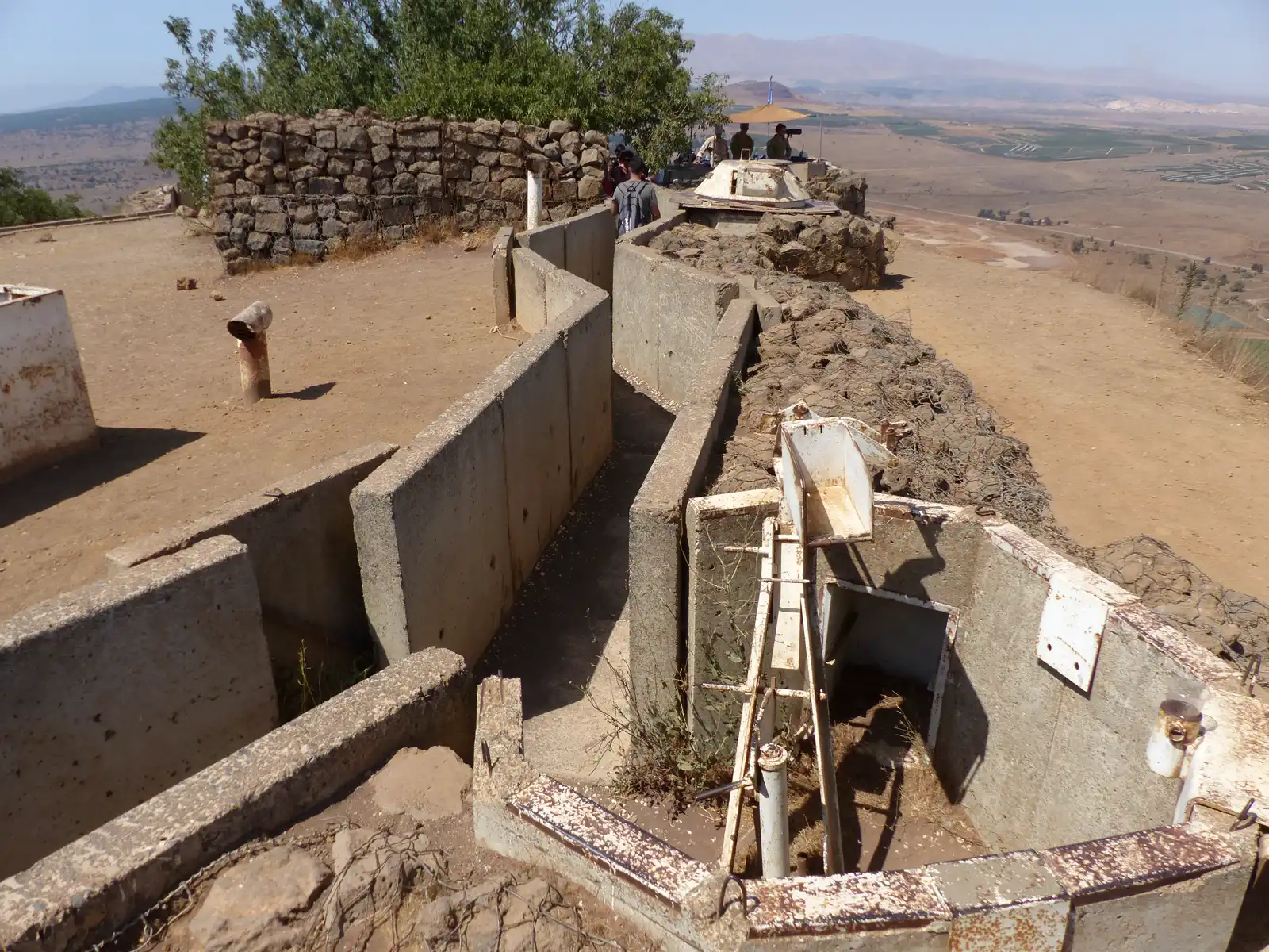 Trincheras y búnkeres de hormigón en una loma pelada de los Altos del Golán, con visitantes asomados a un mirador bajo una sombrilla y una llanura agrícola al fondo.