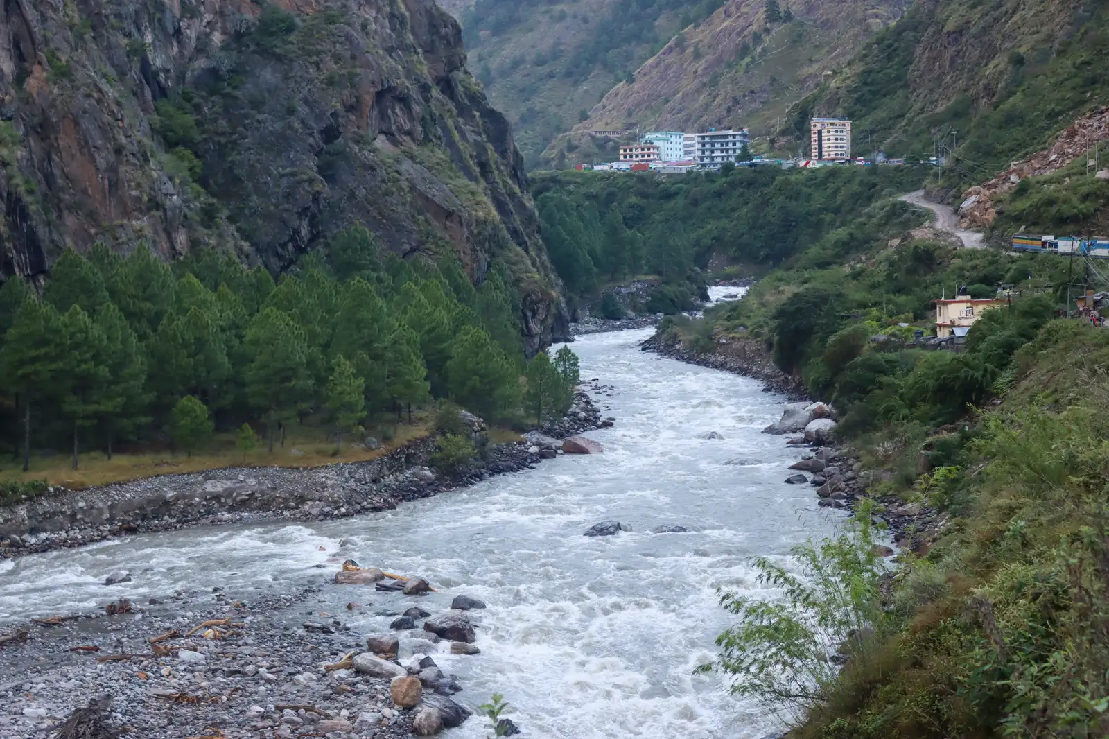 Río caudaloso y blanquecino encajonado entre laderas boscosas y paredes de roca, con un pequeño núcleo de edificios de varias plantas y una carretera de tierra en la ladera derecha