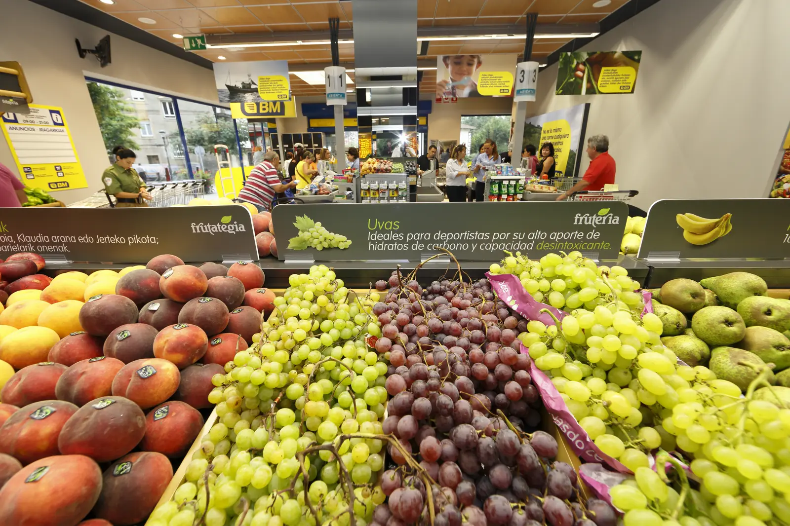Sección de frutería de un supermercado con cajones de melocotones, uvas blancas y negras y peras en primer plano y, al fondo, clientes en las cajas registradoras