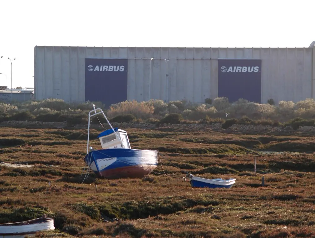 Nave industrial gris con dos grandes carteles azules del logotipo de Airbus, vista desde una marisma con matorral bajo en la que reposan varados dos botes de pesca de madera pintados de azul y rojo.