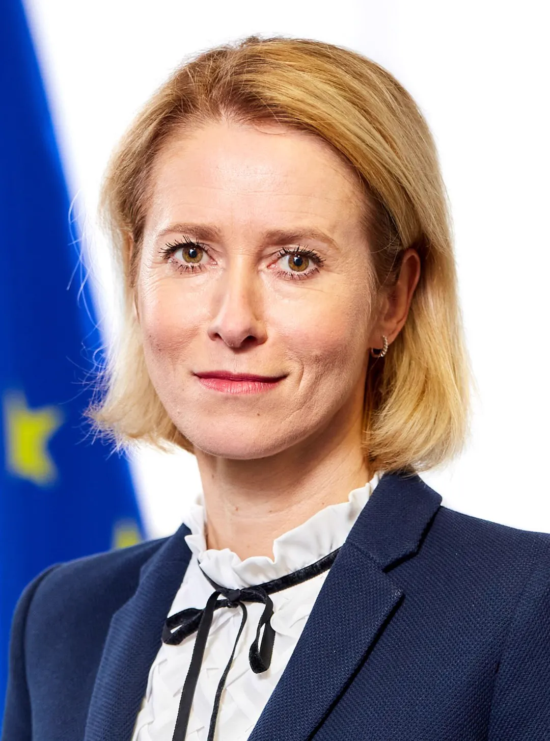 Retrato de Kaja Kallas, de traje azul marino y blusa blanca, ante una bandera de la Unión Europea desenfocada.