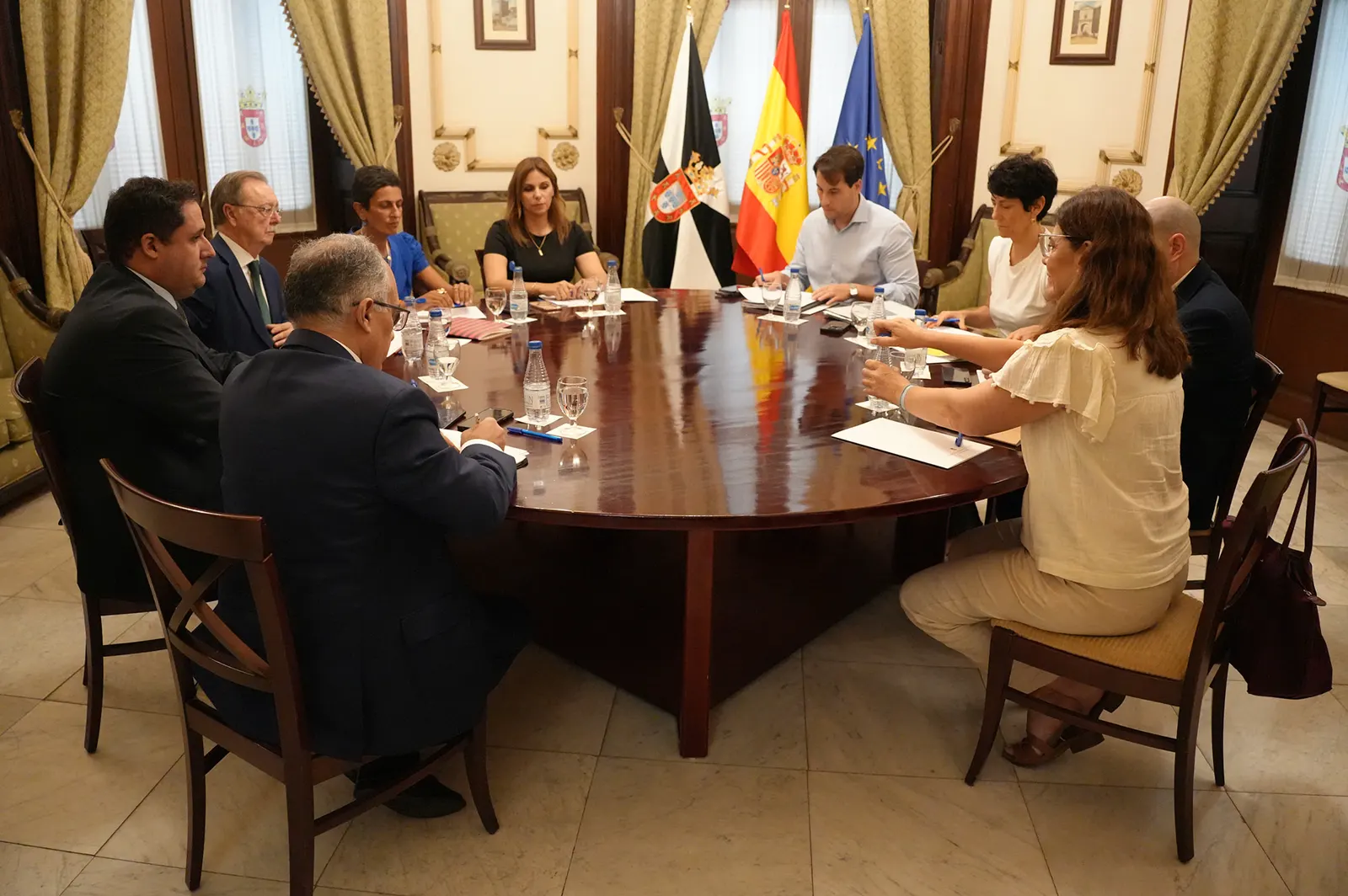 Una decena de personas sentadas alrededor de una gran mesa ovalada de madera en un salón institucional con cortinas verdes; al fondo, las banderas de Ceuta, España y la Unión Europea. Sobre la mesa hay carpetas, botellas de agua y cuadernos.