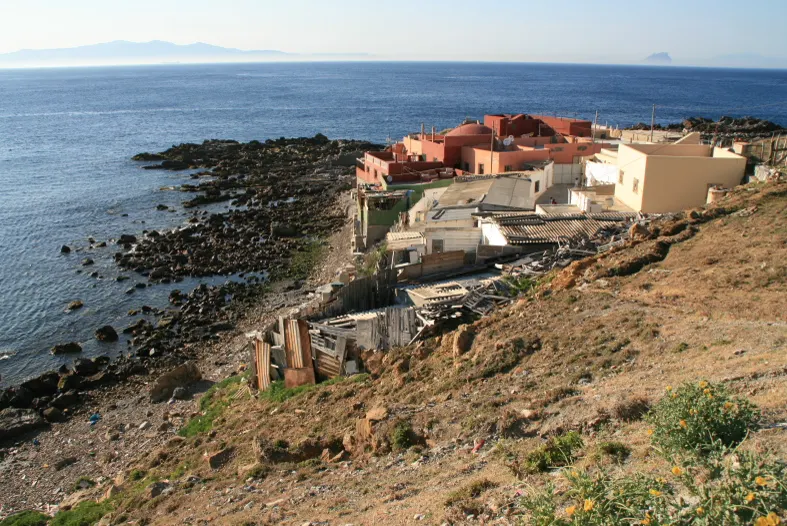 Vista en pendiente de la barriada de Benzú, en Ceuta: casas bajas de colores claros y construcciones de chapa sobre una costa rocosa, con el mar y la costa africana al fondo.