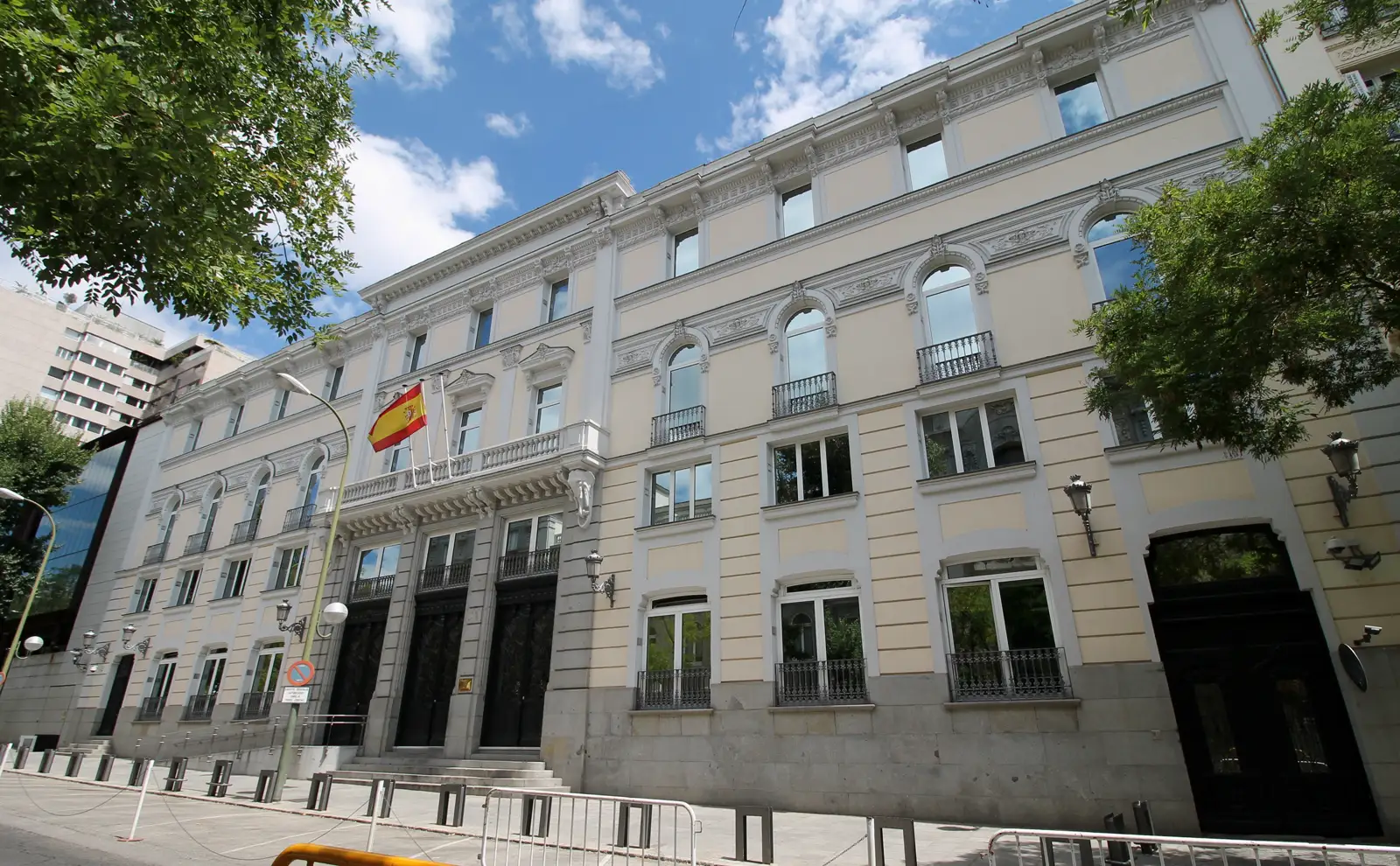 Fachada de piedra clara y tres alturas de la sede del Consejo General del Poder Judicial en Madrid, con ventanas de arco, balcones de forja, faroles metálicos, la bandera de España ondeando sobre la puerta principal y vallas de seguridad en la acera.