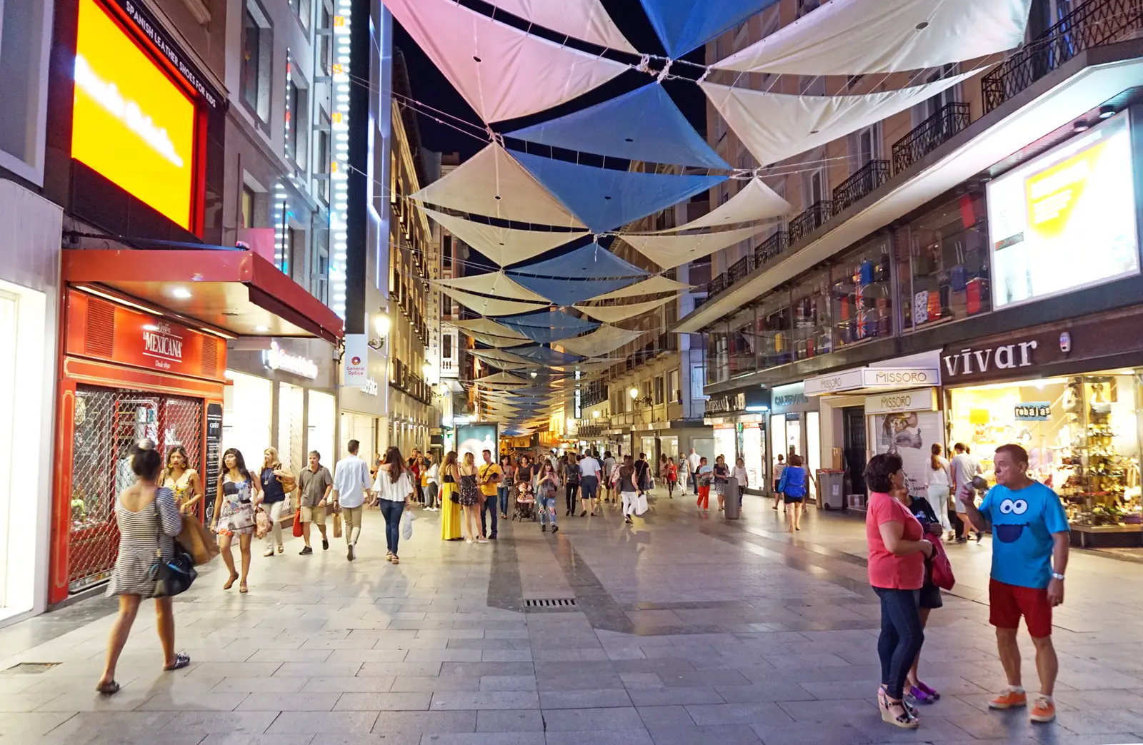Peatones y compradores con bolsas caminan al anochecer por la calle Preciados de Madrid, peatonal y flanqueada por escaparates iluminados, bajo toldos triangulares tendidos entre los edificios.