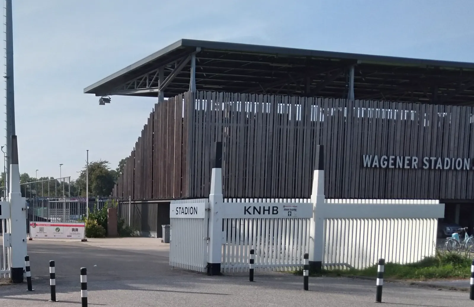 Exterior del Wagener Stadion de Amstelveen: una grada de fachada de listones de madera con el rótulo "Wagener Stadion", una valla blanca con los carteles "Stadion" y "KNHB" y cielo azul al fondo.