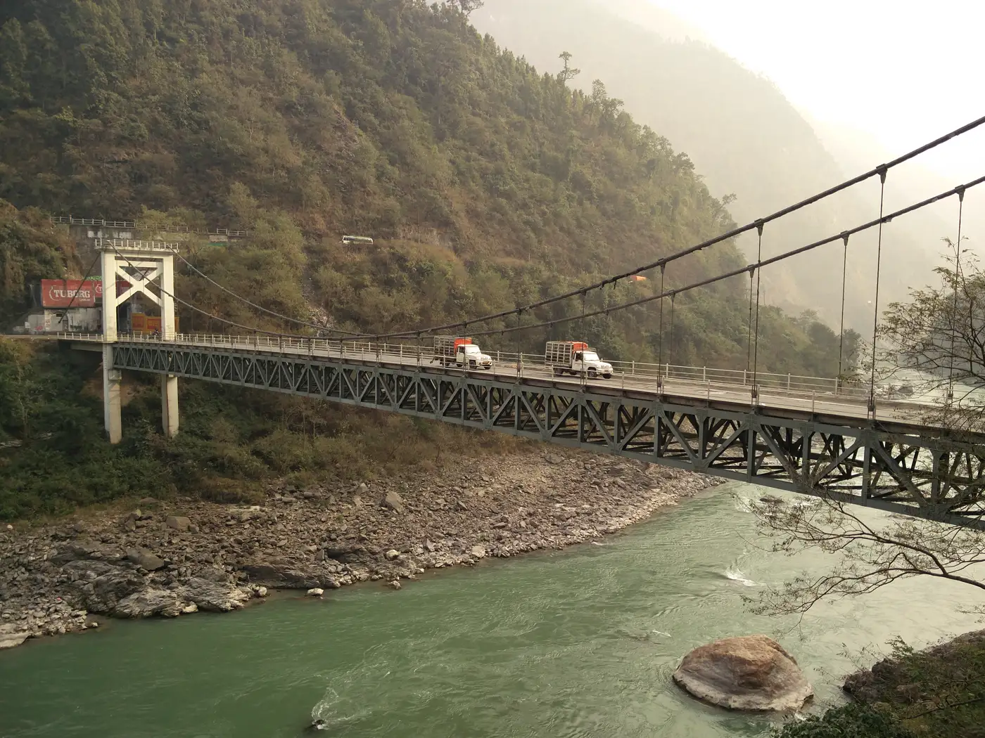 Puente colgante metálico sobre un río de aguas verdosas encajonado entre laderas boscosas, con dos camionetas cruzándolo y una carretera de montaña al fondo