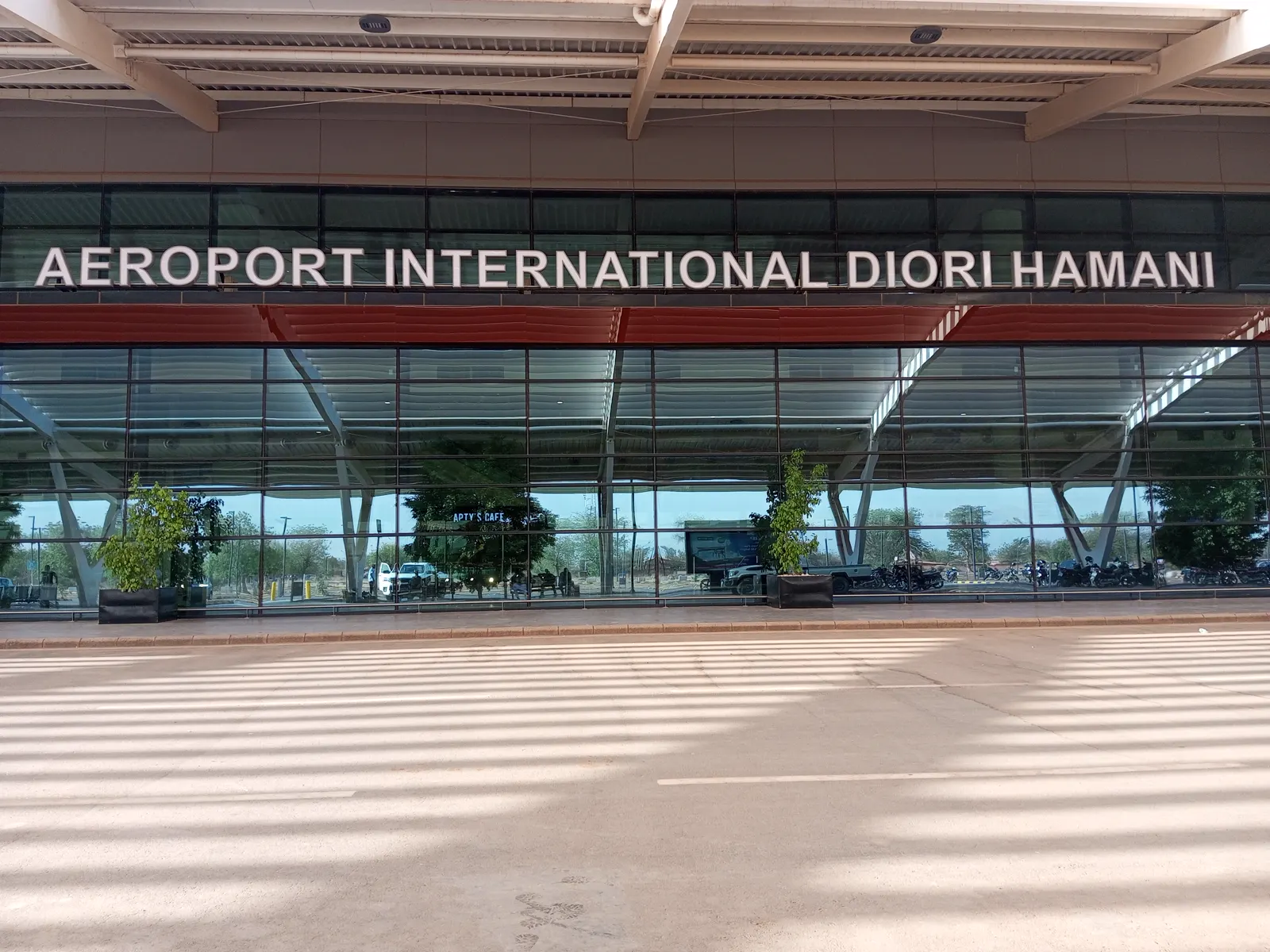 Fachada acristalada de una terminal aeroportuaria con el rótulo «Aéroport International Diori Hamani» sobre los ventanales, una marquesina de vigas metálicas y una acera vacía en primer plano.