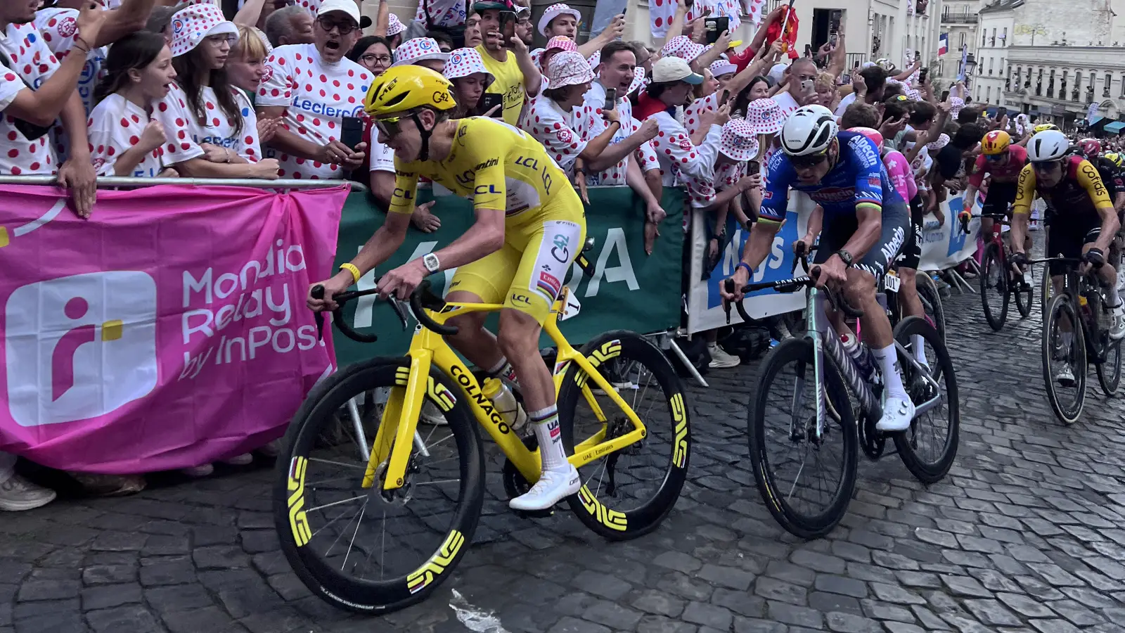 Tadej Pogacar, con maillot y bicicleta amarillos, pedalea sobre adoquines entre vallas repletas de público; detrás de él ruedan otros ciclistas en fila.