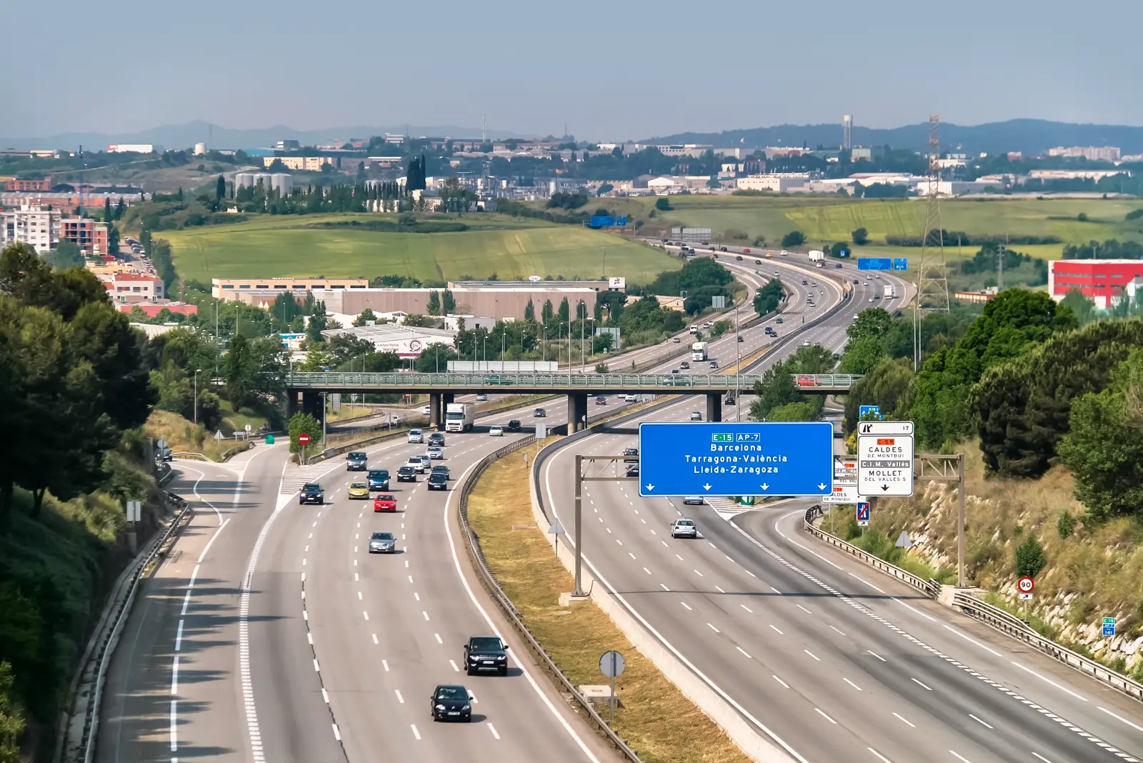 Vista elevada de la autopista AP-7 con tráfico en las dos calzadas y un cartel azul que indica Barcelona, Tarragona-València y Lleida-Zaragoza