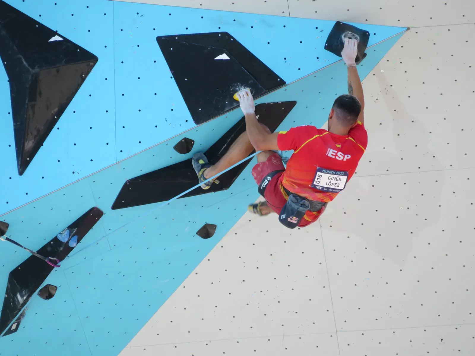 Alberto Ginés López, con la camiseta roja de la selección española y el dorsal 016, escala un muro de dificultad agarrado a una presa negra en un techo inclinado.