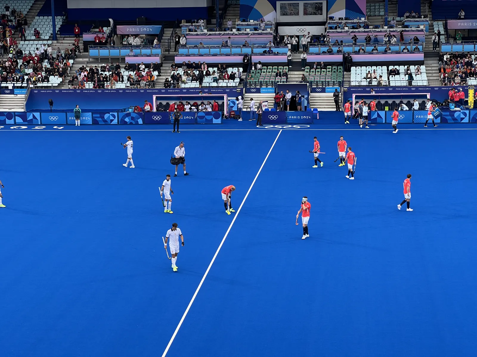 Jugadores de la selección española de hockey hierba, con camiseta roja, y de Gran Bretaña, de blanco, repartidos sobre un campo de césped artificial azul durante un partido de los Juegos Olímpicos de París 2024, con las gradas y las cabinas de prensa al fondo.