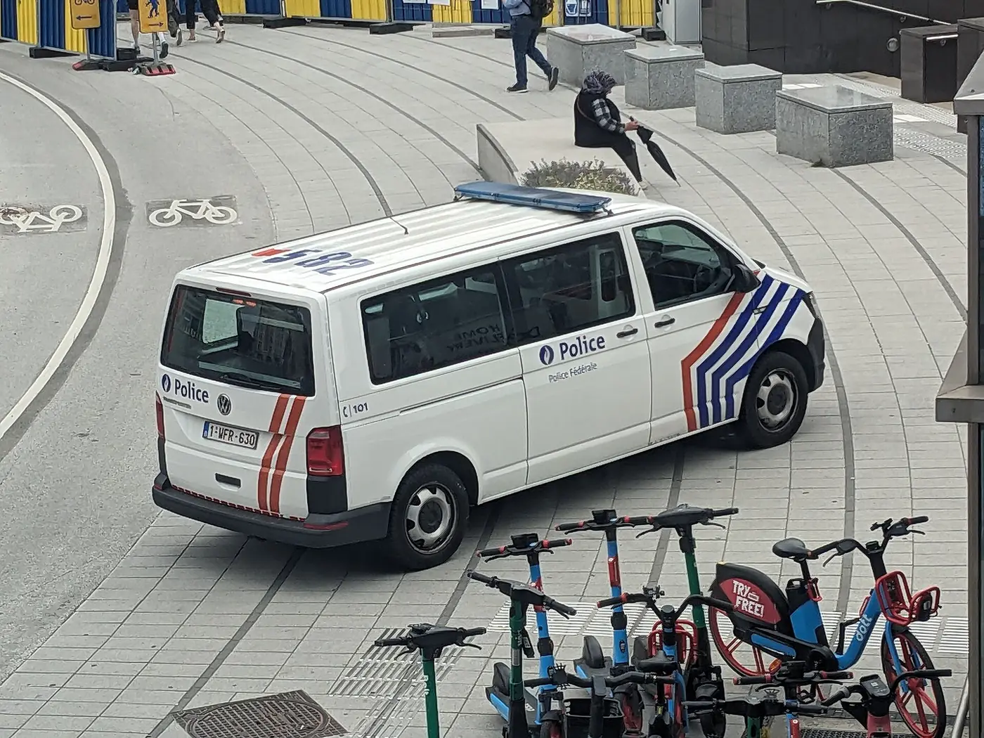 Furgón Volkswagen Transporter de la Policía Federal belga, blanco con franjas azules y amarillas y rotulación «Politie / Police», aparcado en una plaza de Bruselas con edificios de oficinas al fondo.